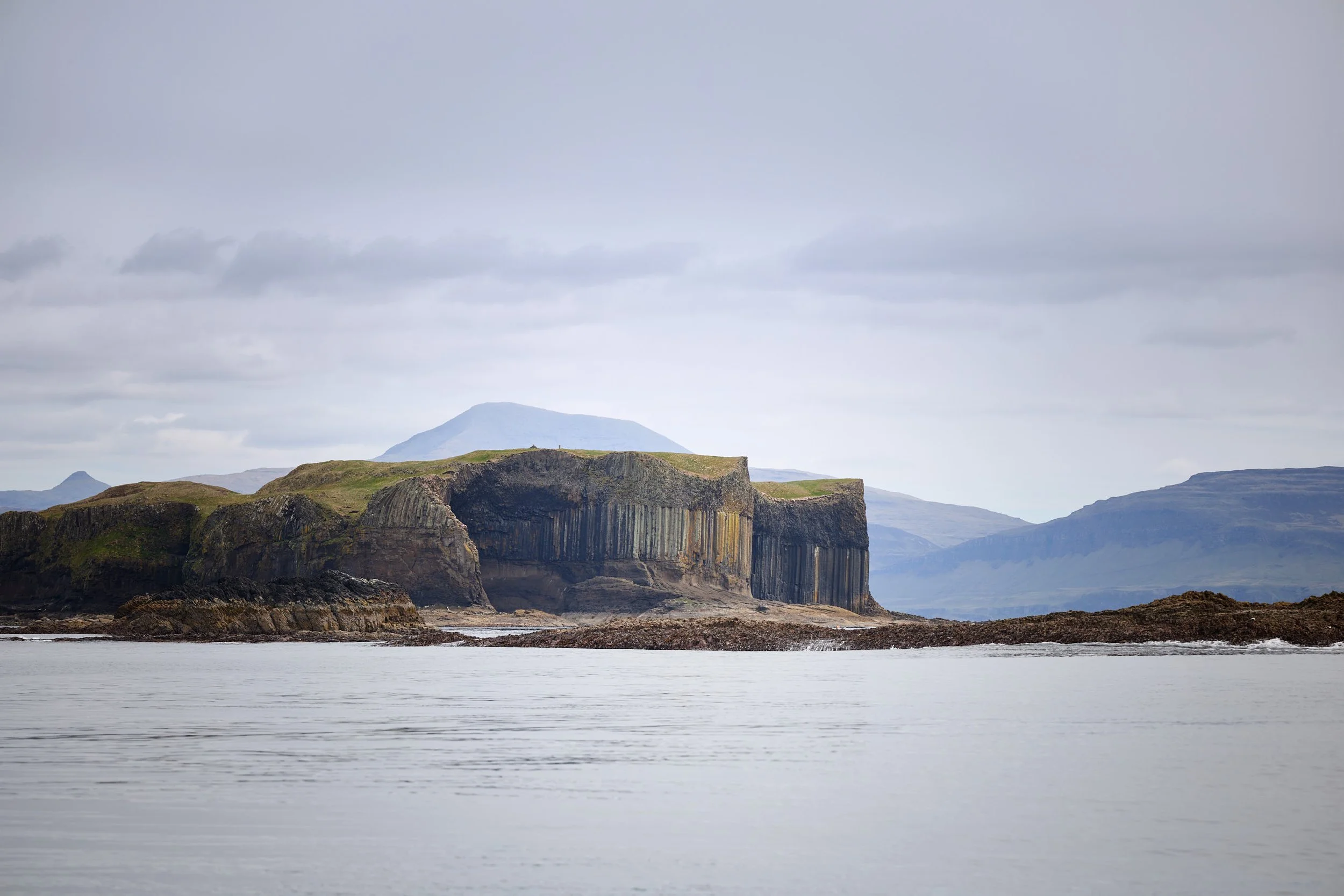 Coastal landscape with sea, steep cliffs, and mountainous terrain in the background under a cloudy sky.