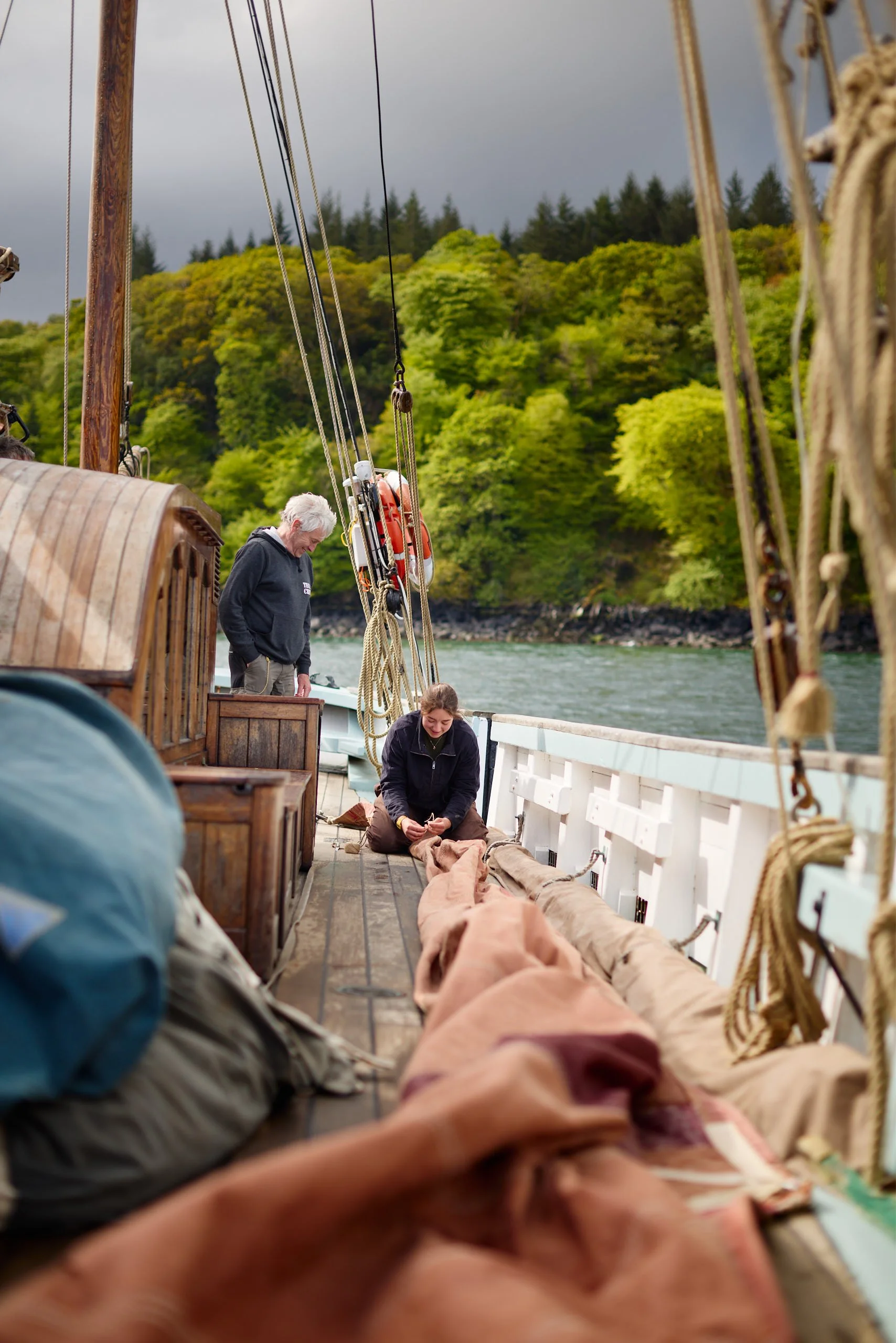 People sitting and standing on the deck of a boat, with a background of lush green trees and cloudy sky.