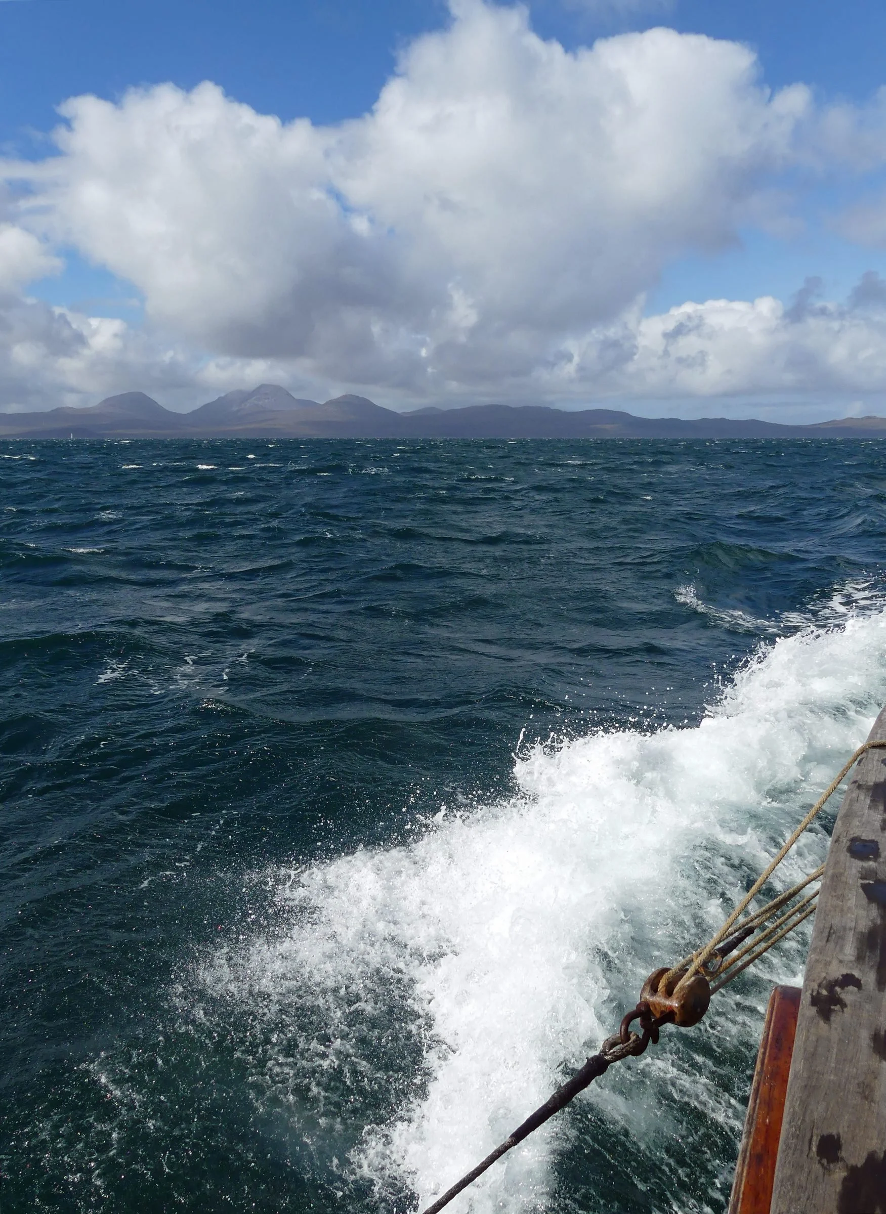 A view from a boat on the open ocean, with dark blue water and white wakes, with distant mountains and partly cloudy sky.