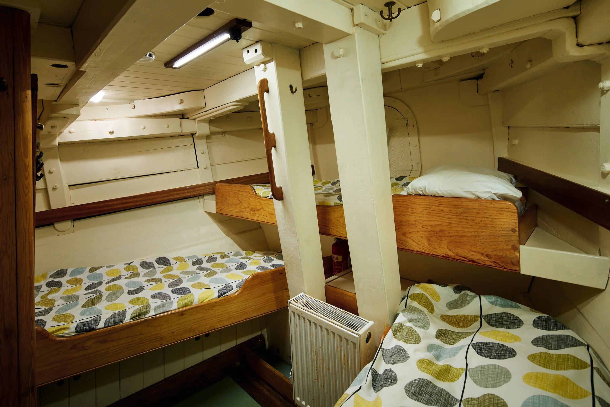 Interior of a vintage wooden train compartment with three beds, each with patterned bedding; one bed has a pillow. There is a radiator heater below the beds and a fire extinguisher on the wall. The ceiling has a fluorescent light and wooden beams, an