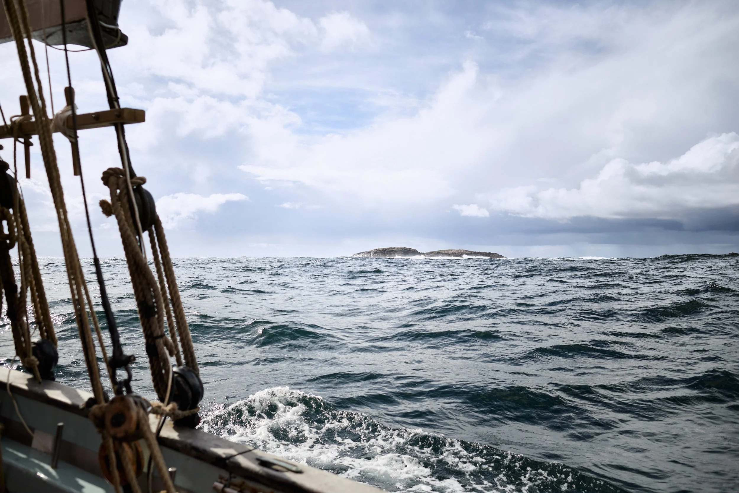 View of the ocean as seen from a sailing boat, with ropes and rigging on the left and a small island in the distance under a partly cloudy sky.