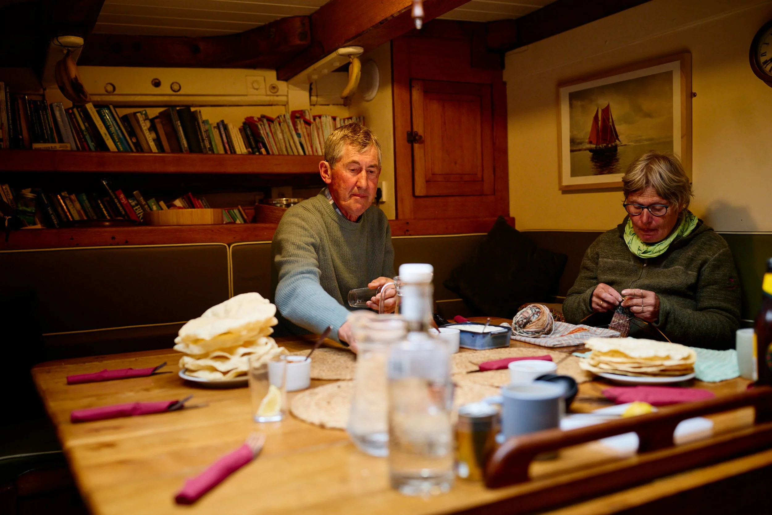 Two older men are seated at a wooden table engaged in knitting or other needlework. The table is scattered with yarn, knitting needles, and other crafting supplies. The background features a wall with a large bookshelf filled with books, a painting of a sailboat, and wooden cabinets. The room has warm lighting and a cozy ambiance.