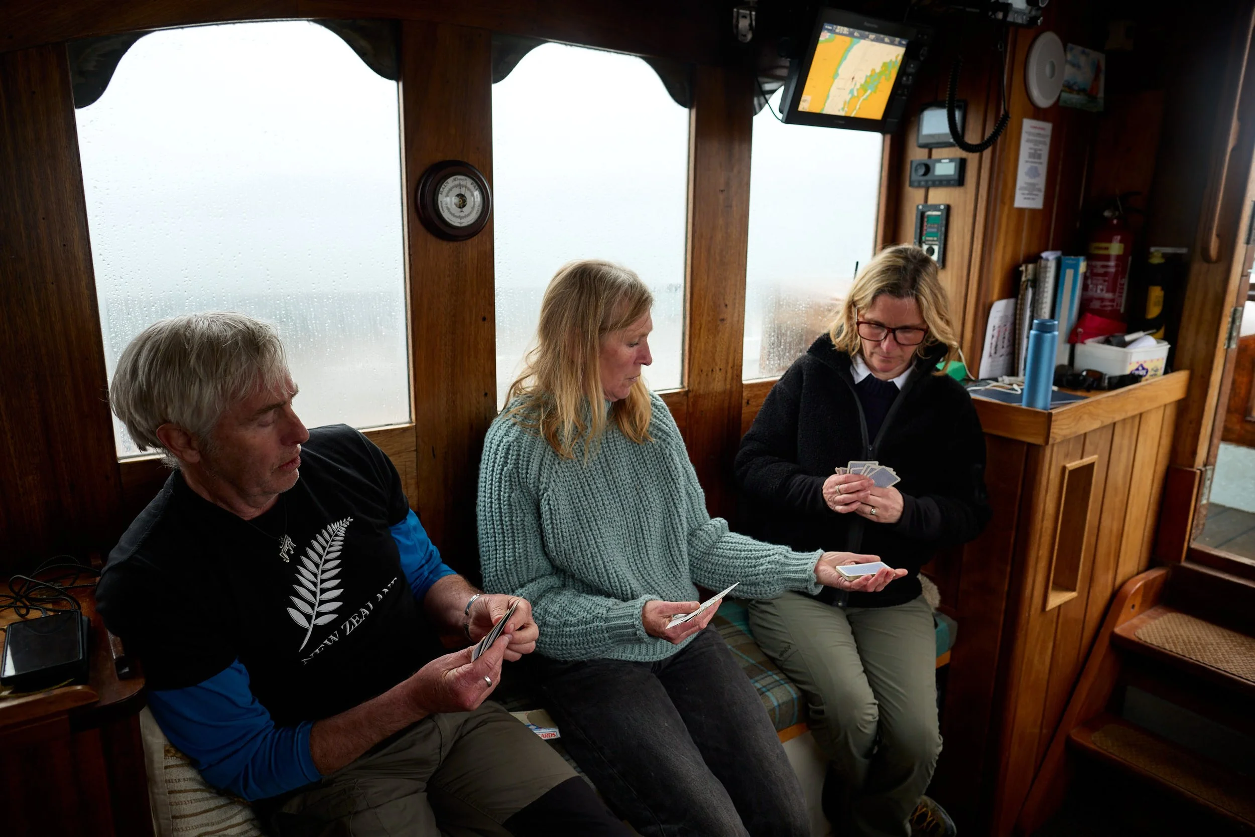 Three people sitting in a boat's cabin, playing cards and looking at their phones. The cabin has wooden walls, windows with rain and a navigation screen displaying a map.