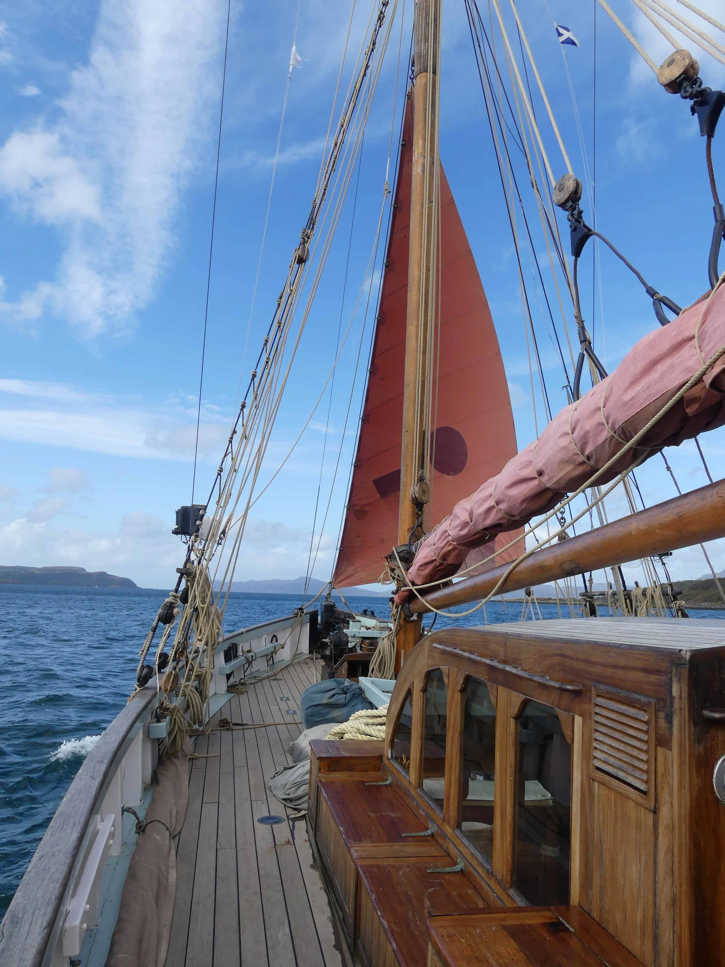 View from a sailboat with wooden deck and gear, orange sail, blue sky with clouds, distant land on the horizon.
