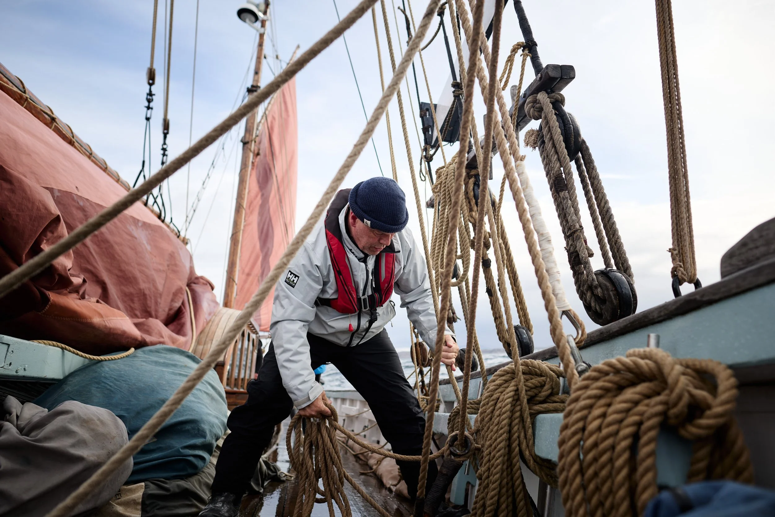 A person working on a sailing ship, handling ropes among various rigging and equipment on deck with others working around.