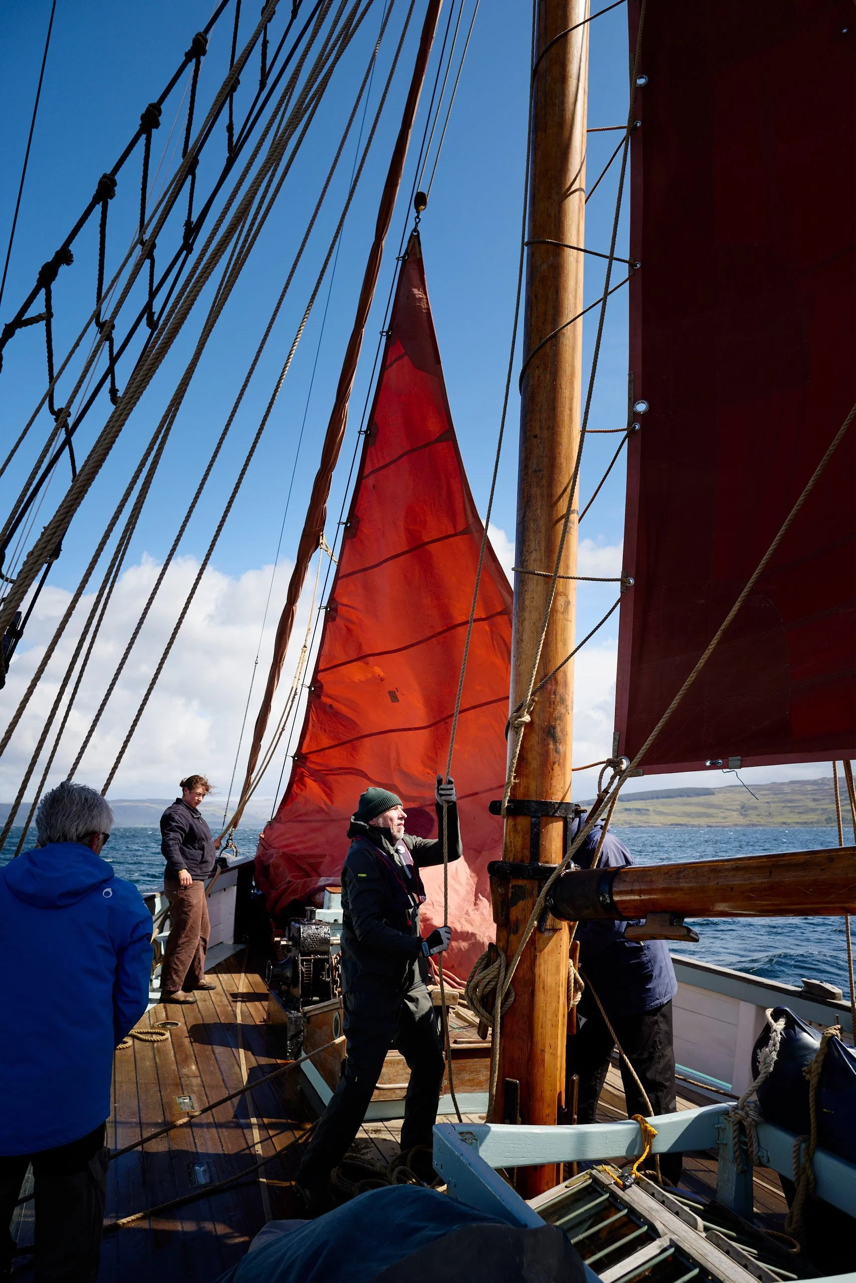 People sailing on a wooden sailboat with a large red sail, several crew members are working with ropes and equipment on the deck, blue sky and water in the background.
