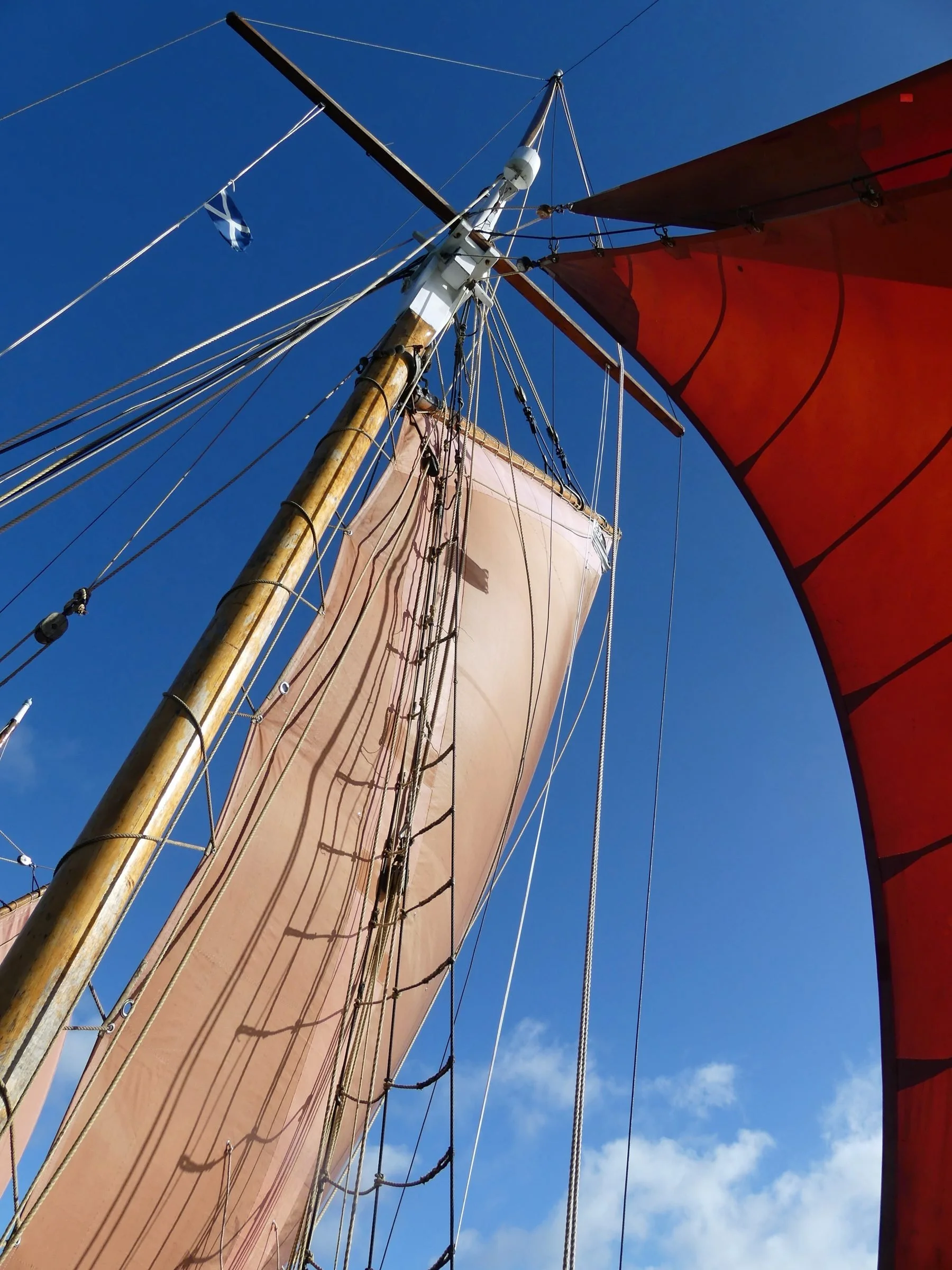 Looking up at a tall ship's mast with beige sails, red sails, and numerous ropes against a clear blue sky with a few clouds.
