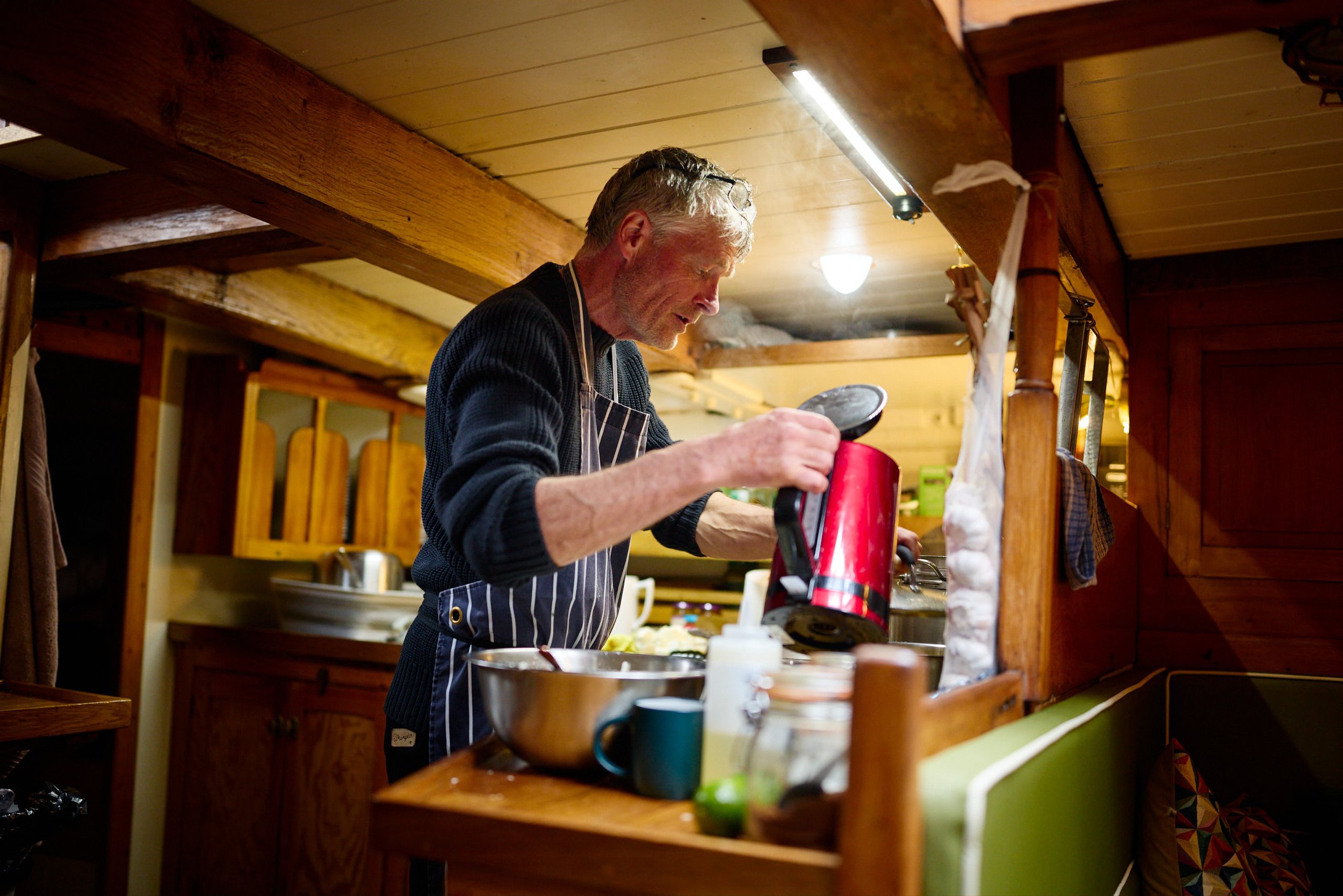 An older man with gray hair working in a cozy wood-paneled kitchen, pouring hot water from a red thermos into a mug while wearing glasses on his head and a black sweater, surrounded by kitchen items and utensils.