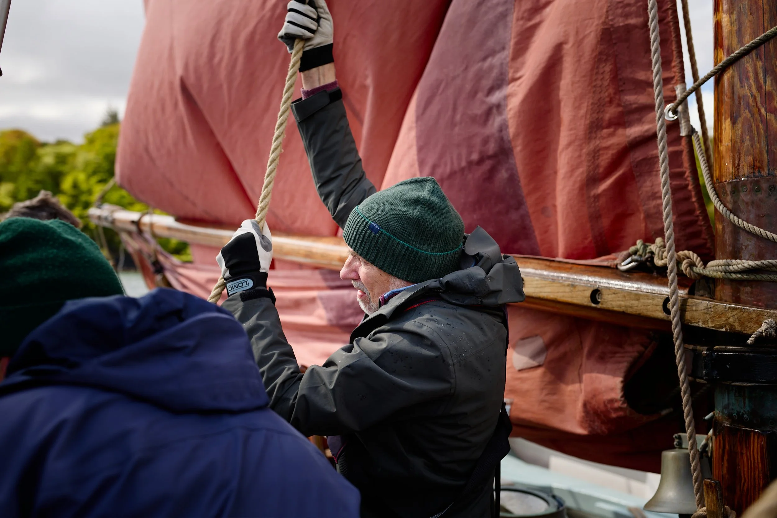 A group of people working on the rigging of a sailboat, with one person securing a rope while others observe, against a background of cloudy sky and lush greenery.