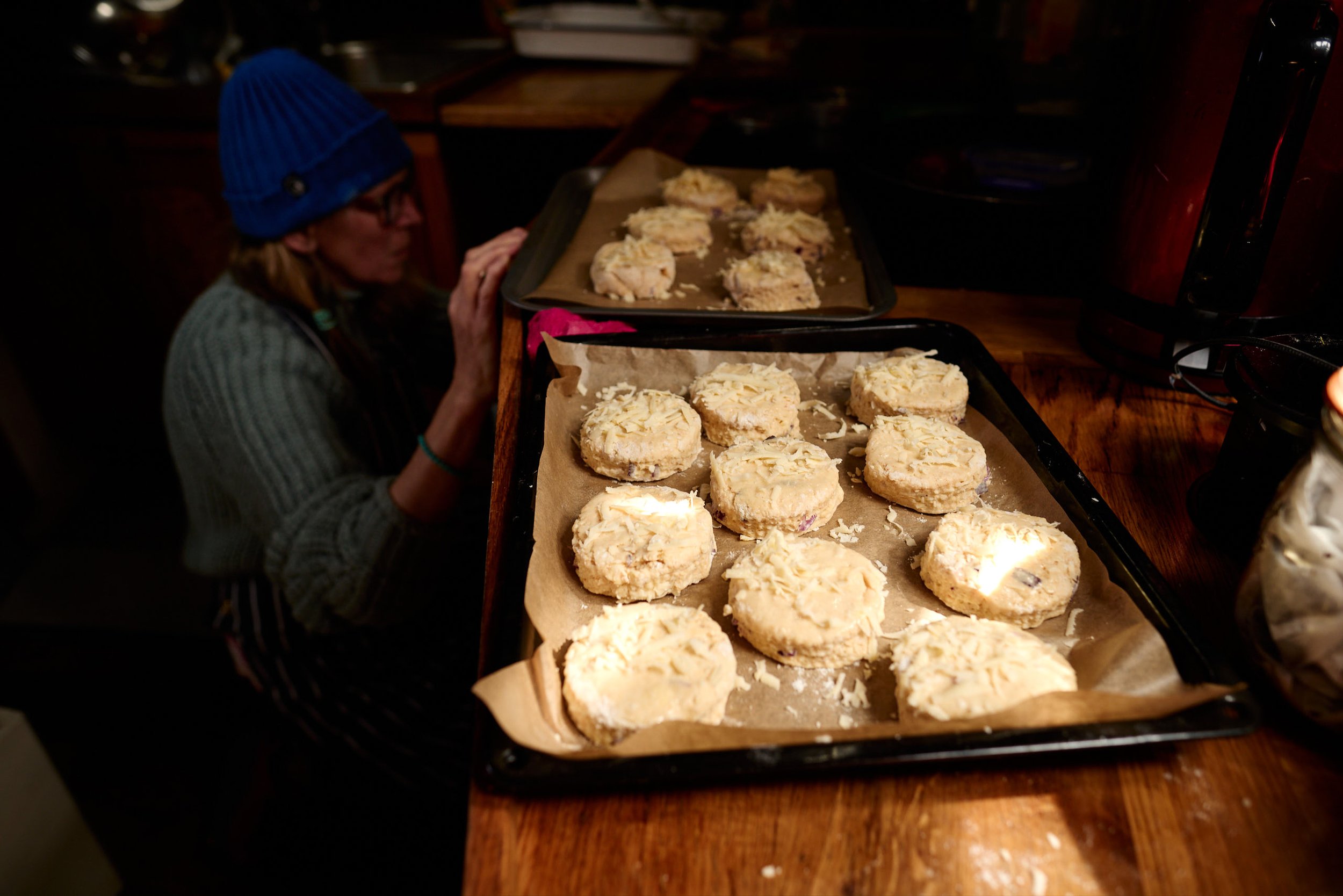 Close-up of a baking sheet filled with glazed cookies topped with shredded white chocolate, with a person wearing a blue beanie and glasses in the background, sitting at a wooden table in a dimly lit kitchen.
