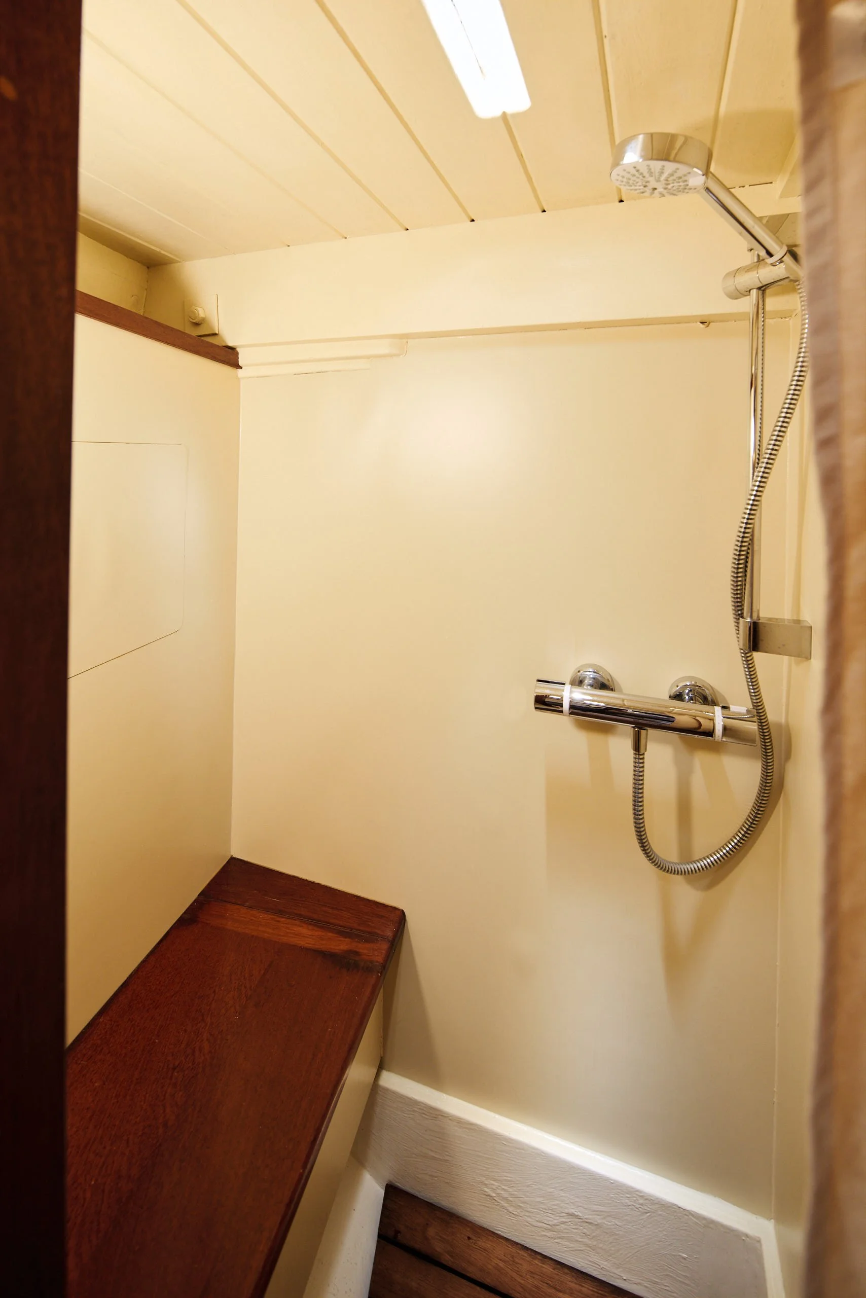 View of a walk-in shower with a chrome handheld showerhead, a dark wooden seat, beige walls, and a white ceiling with a fluorescent light.