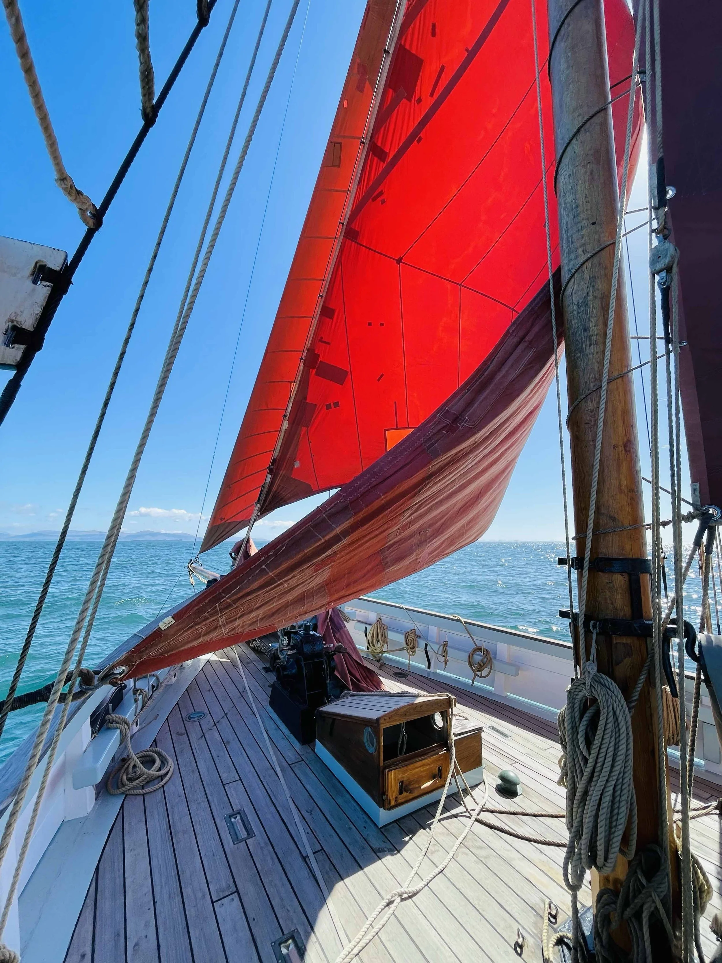 View of a sailboat deck with a large, vibrant red sail, rigging ropes, and wooden fixtures. The background shows blue sky and ocean with distant landforms.