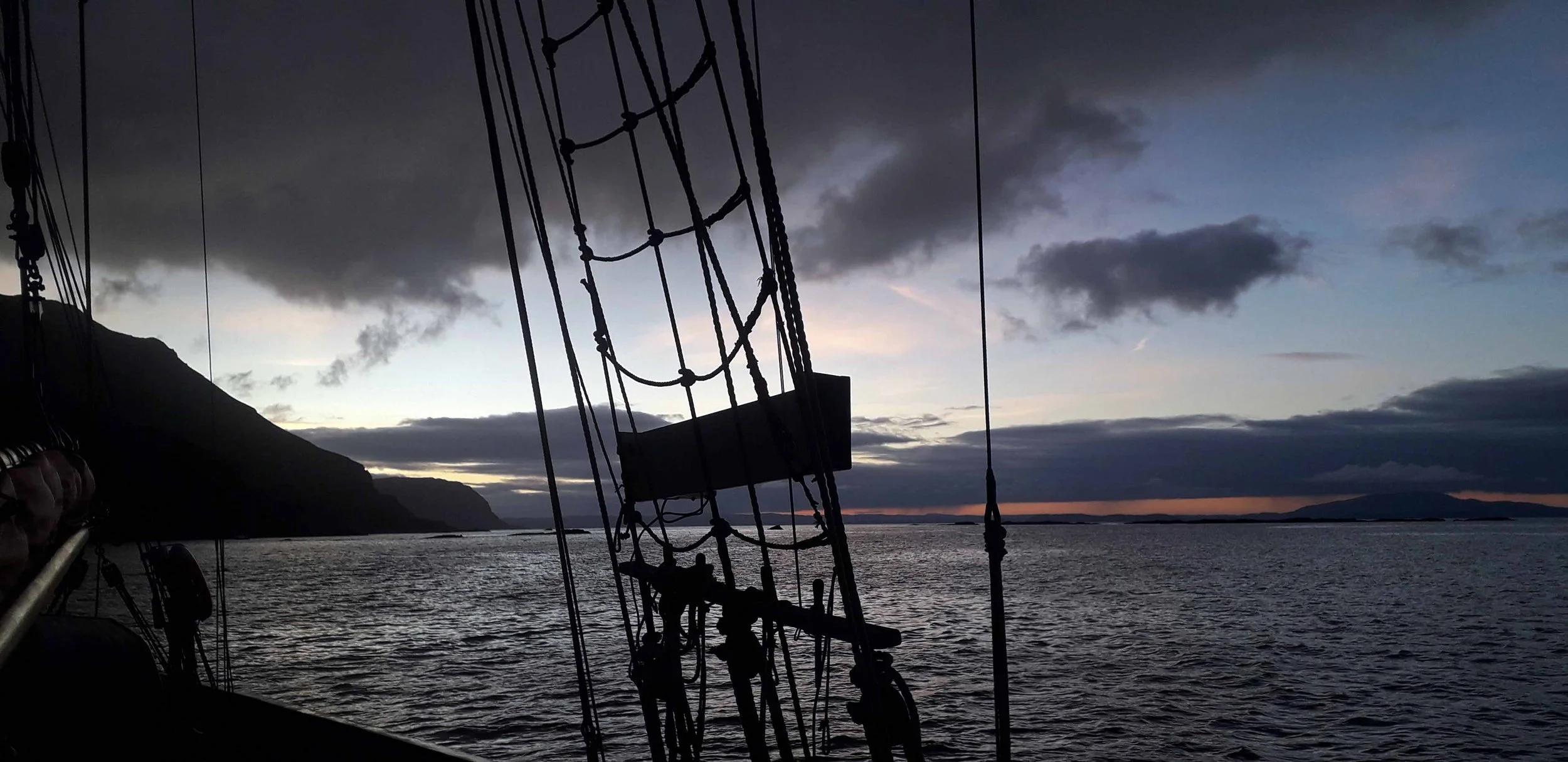 A view from a sailing ship at sunset or dusk, with the ship's rigging and masts silhouetted against a cloudy sky over calm water, with distant landforms on the horizon.
