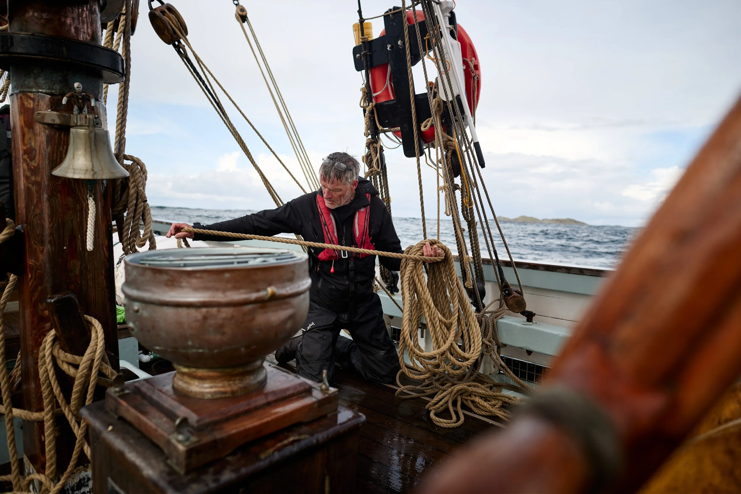 Steve Jones, Skipper of Provident  on a sailing ship adjusting ropes amid an array of ropes, pulleys, and nautical equipment, with the ocean and cloudy sky in the background.