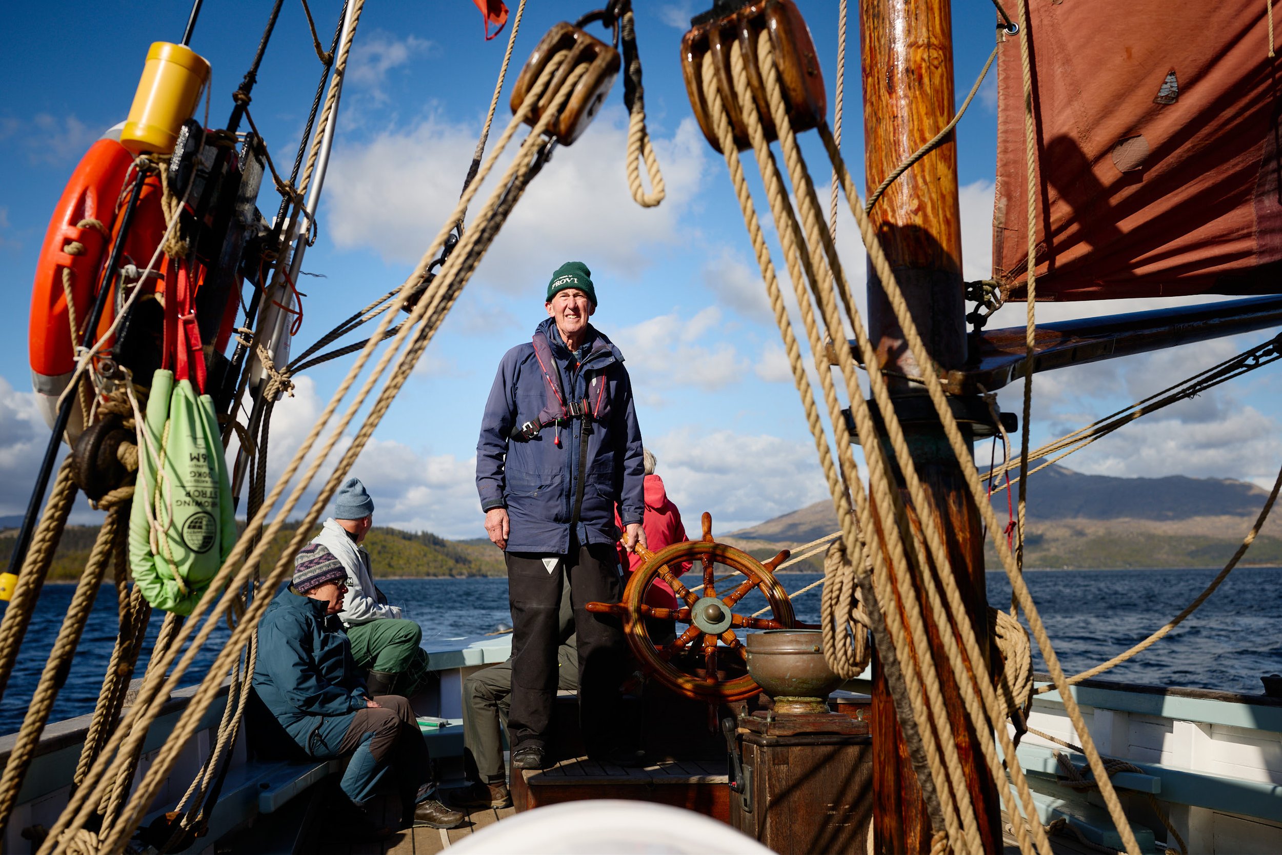 People on a sailing ship with a man standing at the helm, surrounded by ropes, with the blue sky and water in the background.
