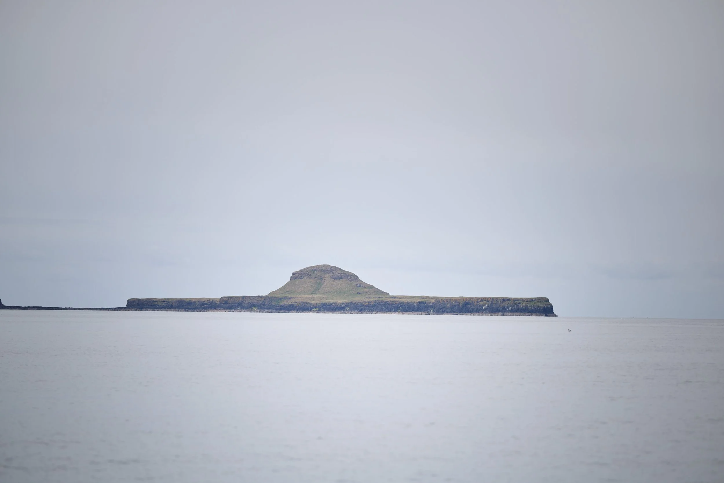 A large, flat island with a small hill in the middle, surrounded by water under a cloudy sky.