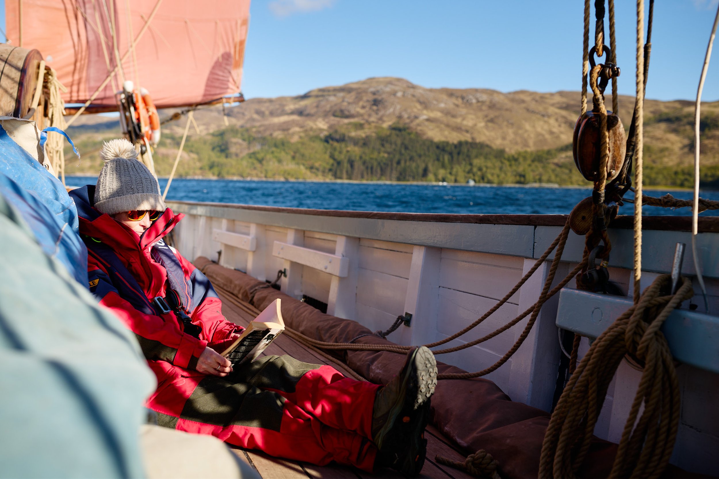 A person in a red jacket, gray knit hat, and sunglasses sitting on a boat, reading a book with scenic mountains and water in the background.