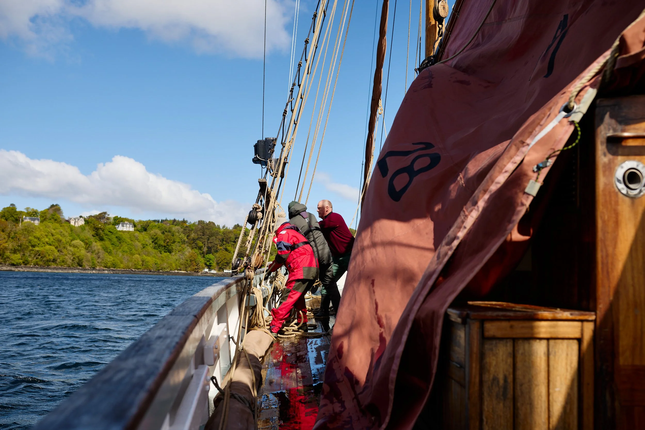 People aboard a wooden sailing boat, tied to the rigging with a scenic shoreline with trees and houses in the background.