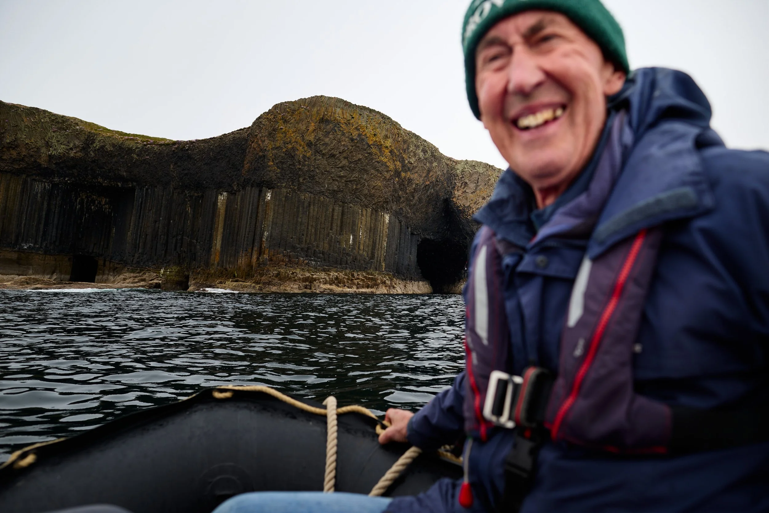A man in a green beanie and blue jacket paddling a kayak on water near Fingal's Cave  with tall columns and green moss, smiling with teeth showing.