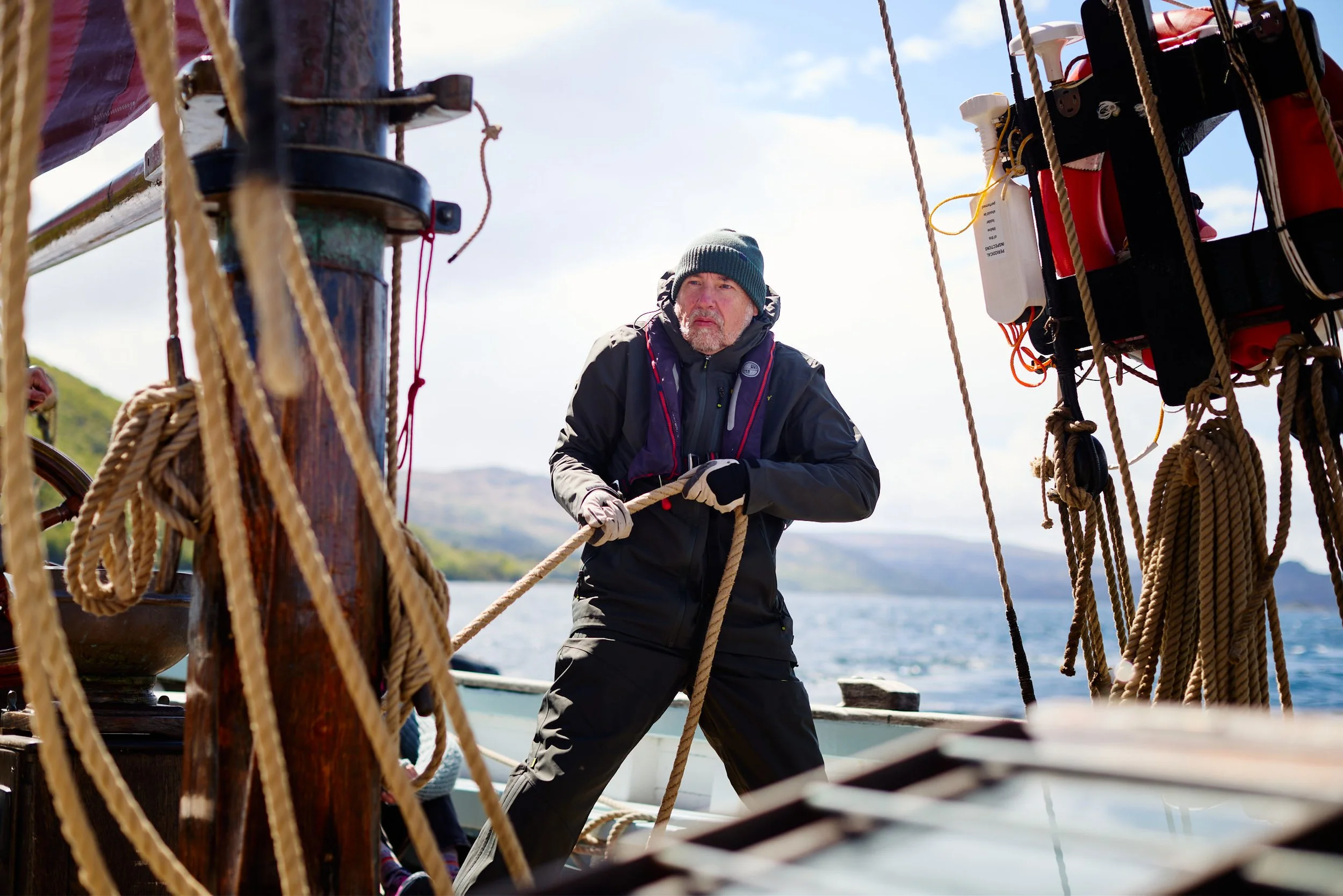 A man wearing a gray beanie and black jacket holding onto ropes on a sailing ship, with a background of water and sky.