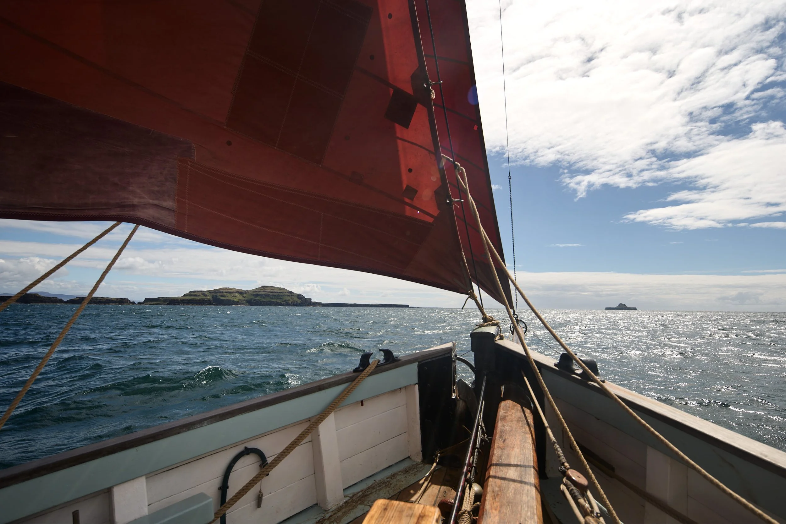 A view from inside a boat looking forward at the red sail and the ocean with an island in the background under a partly cloudy sky.