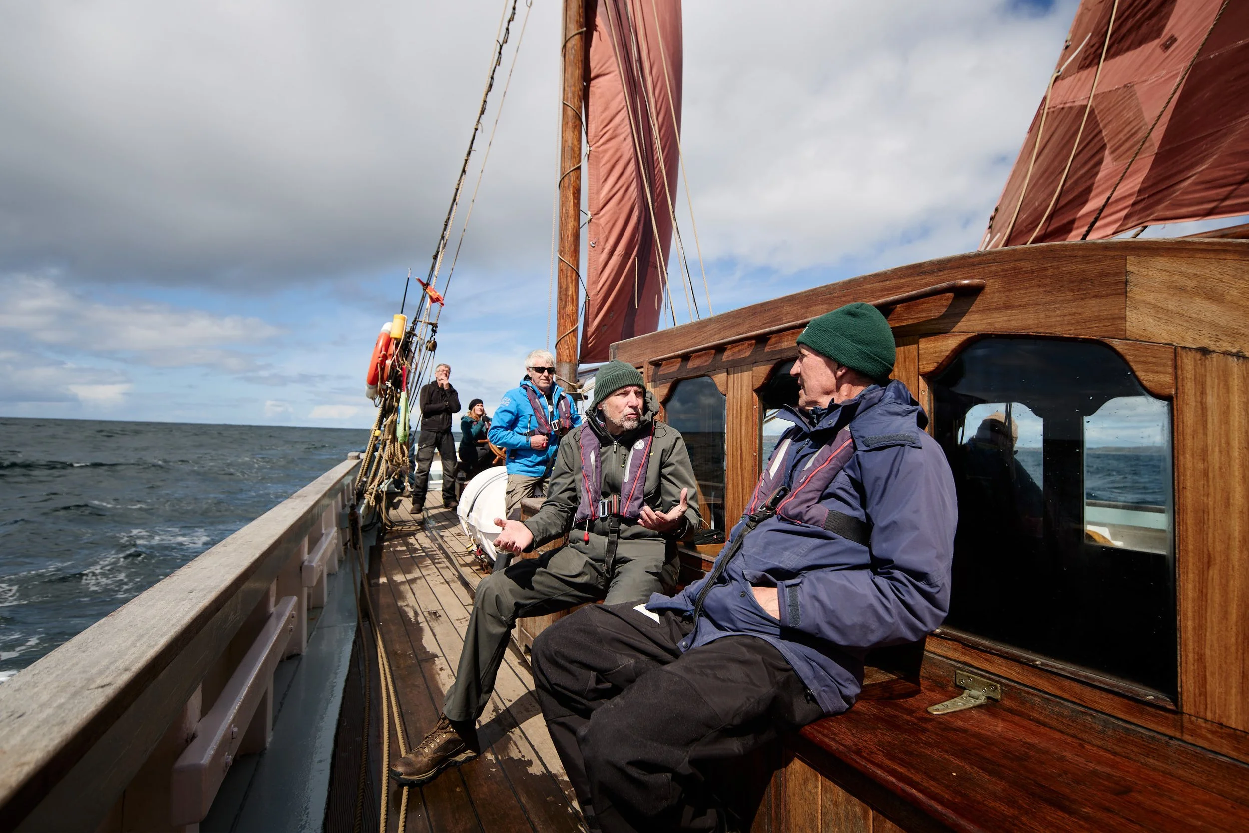 A group of people on a wooden sailboat with red sails, sailing on open water under a partly cloudy sky, some wearing jackets and hats.