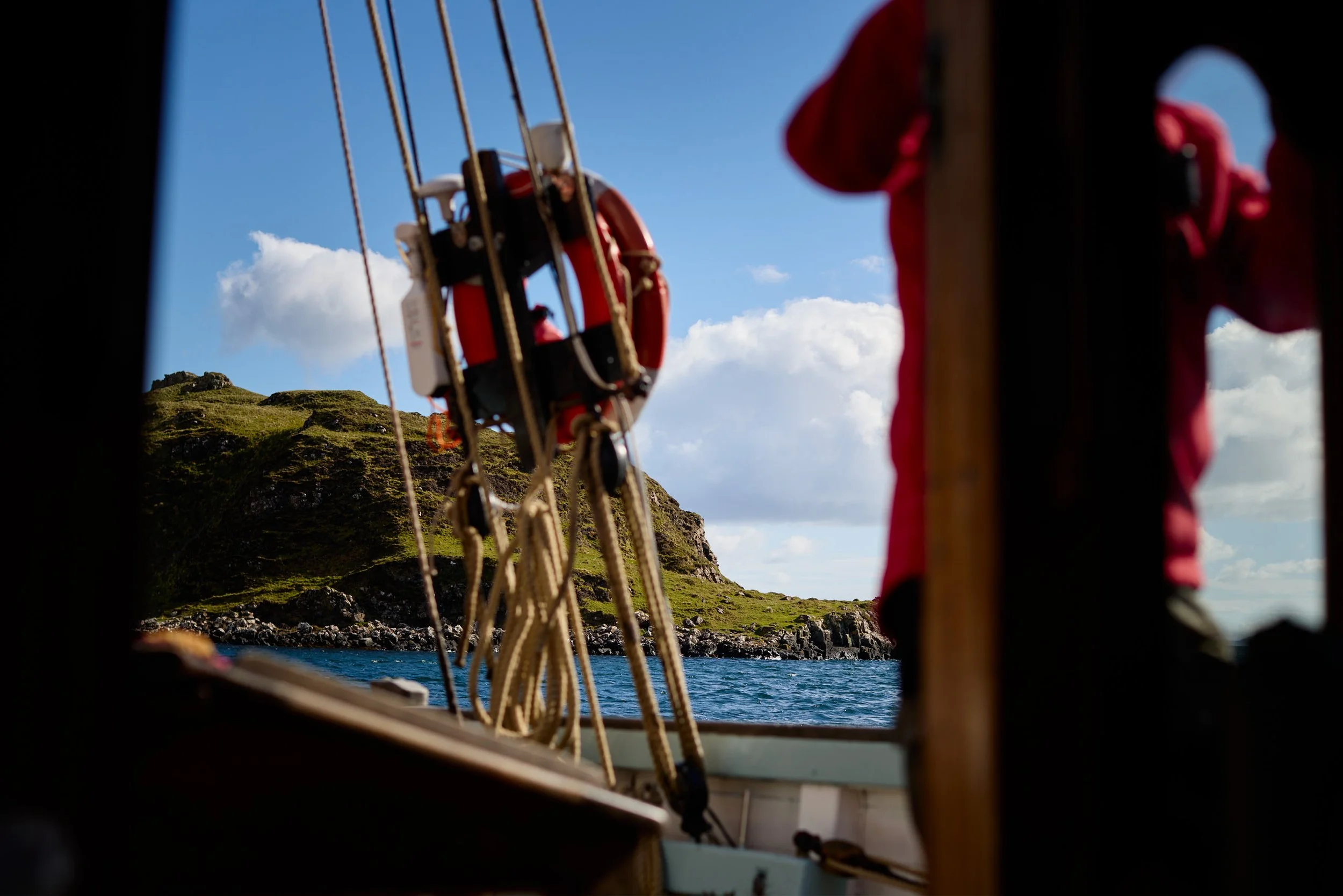 View of a coastal landscape through the window of a boat, with green hills, rocks, and blue sky with clouds in the background.