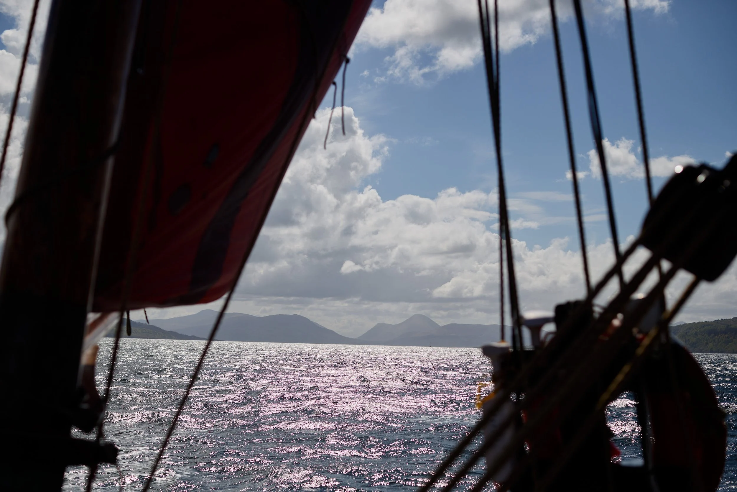 View of a water body with mountains in the distance, captured from a boat with part of the boat framing the image in the foreground, along with some rigging and a sail, under a partly cloudy sky.
