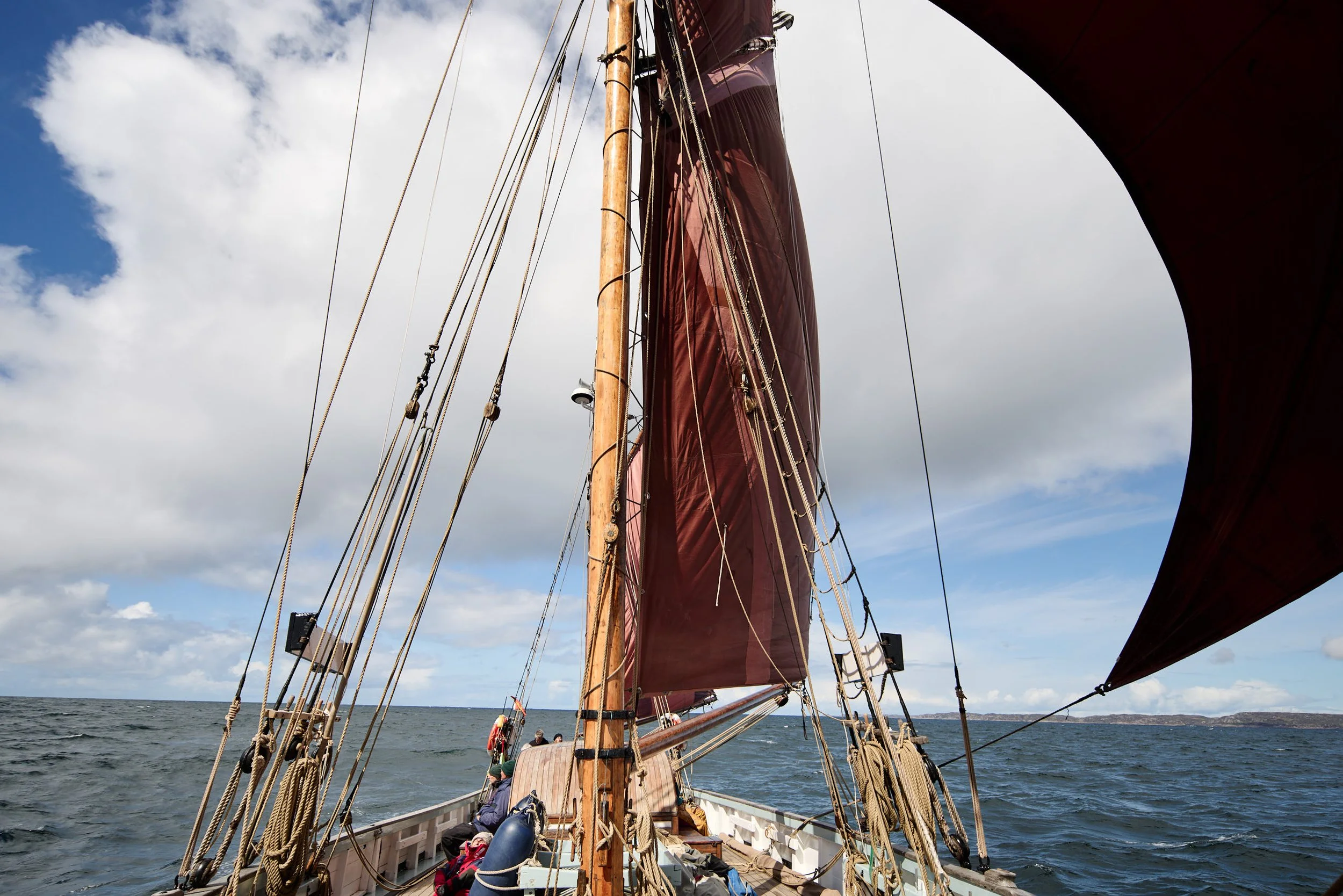 A view from the deck of a sailing ship, Provident,  looking towards the mast and sails, with people seated on the deck, on open water under a partly cloudy sky.
