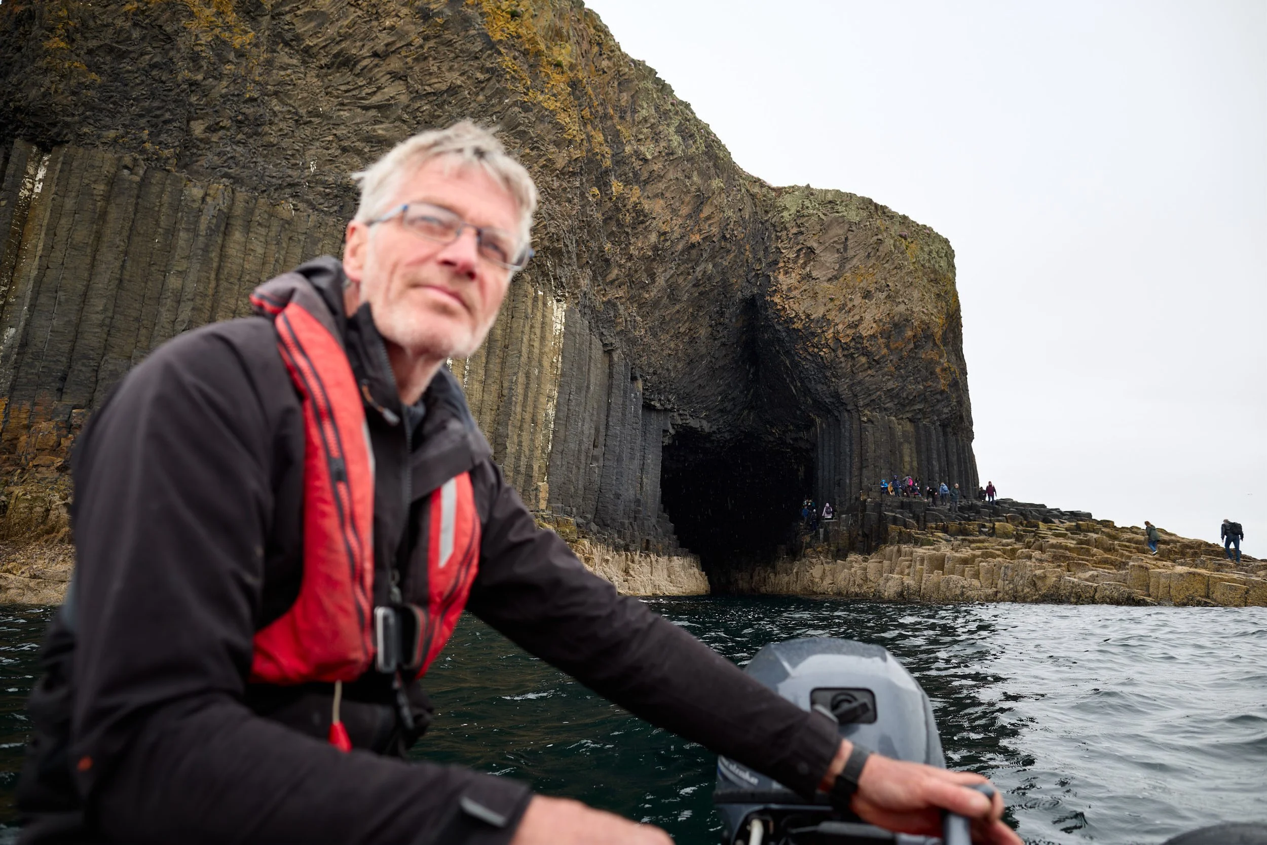 A man on a boat near a large sea cave with people walking along the rocky shoreline.