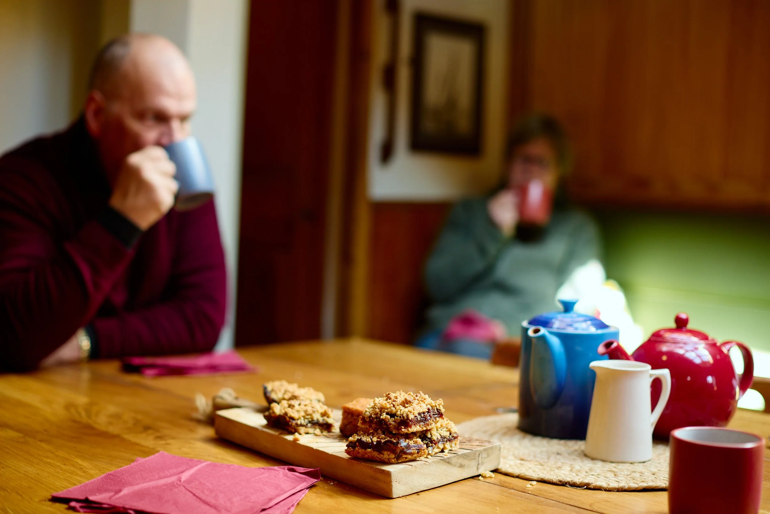 People sitting at a table with tea cups enjoying a snack, with plates of cookies and a wooden board of food on the table.