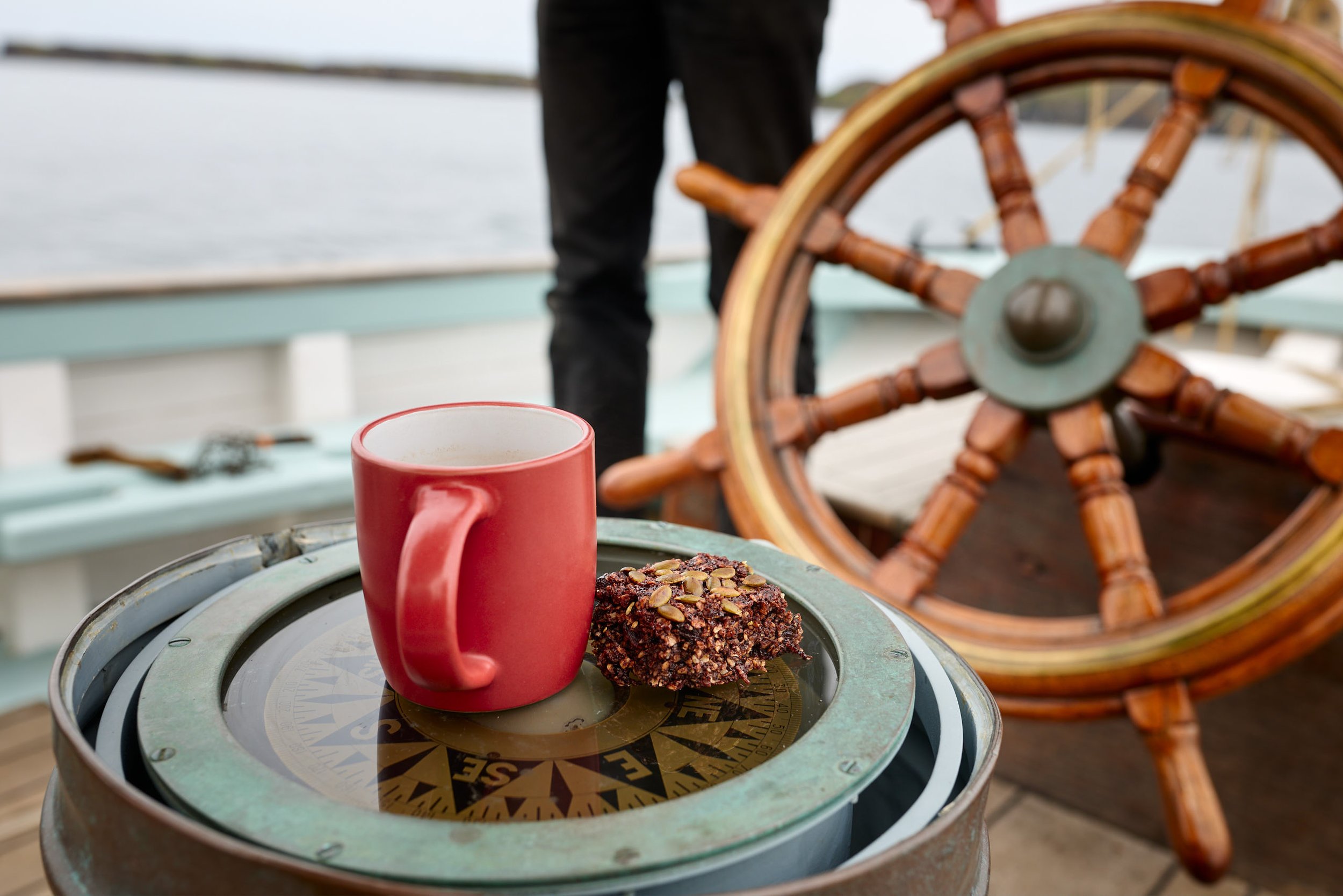 A red mug, a dessert topped with pumpkin seeds, and an orange tabby cat lying nearby, with a ship's wheel and a compass on a table on a boat.