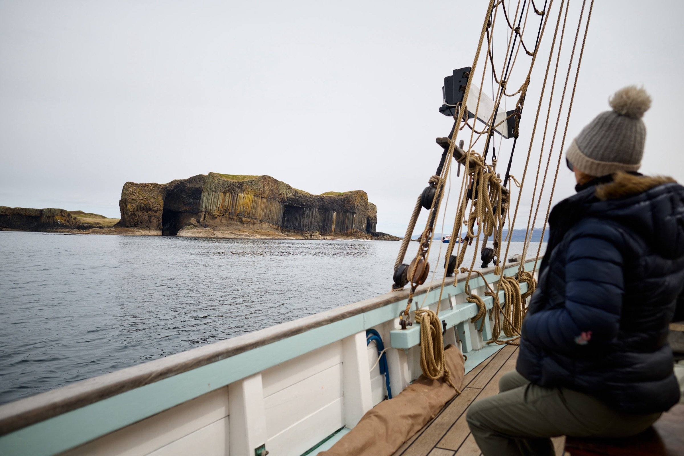 A person wearing a knit hat and dark jacket sitting on the deck of Provident, looking towards a Scottish island with grassy cliffs and rock formations, with calm water around.