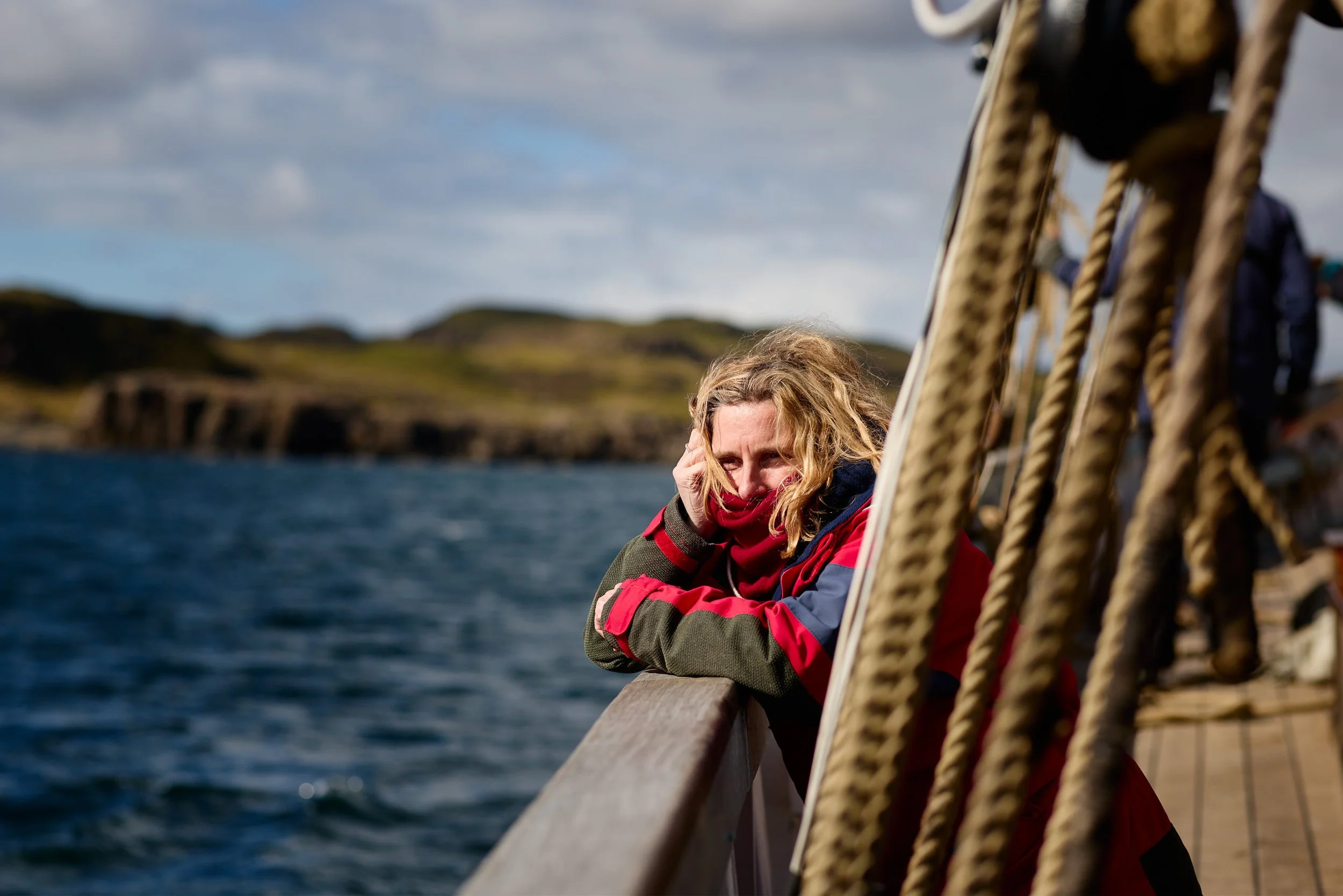 A woman in a red and gray jacket sitting on a boat railing, with a scenic coastal background and cloudy sky.