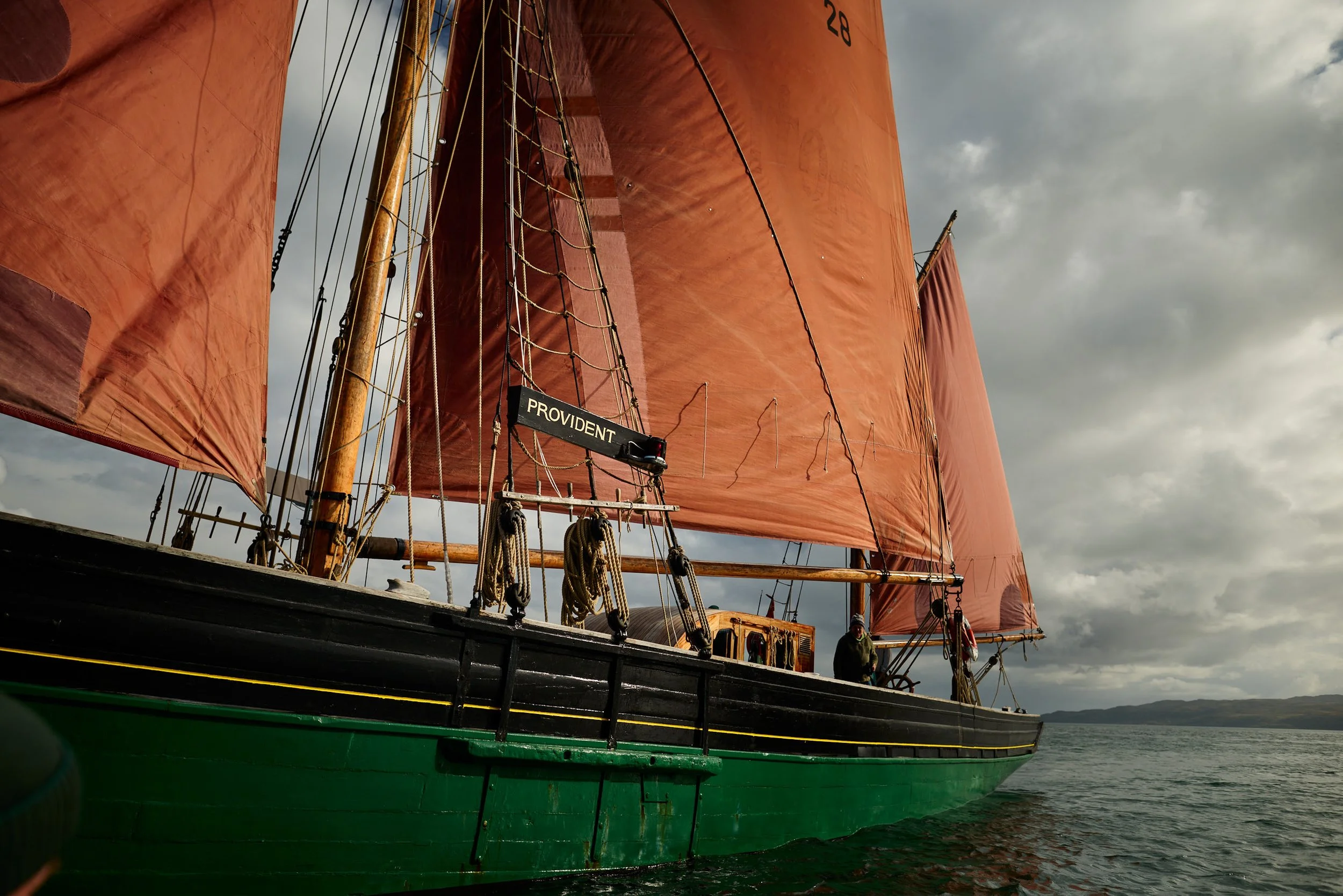 A sailing ship with orange sails and a green hull, on water with a cloudy sky in the background.