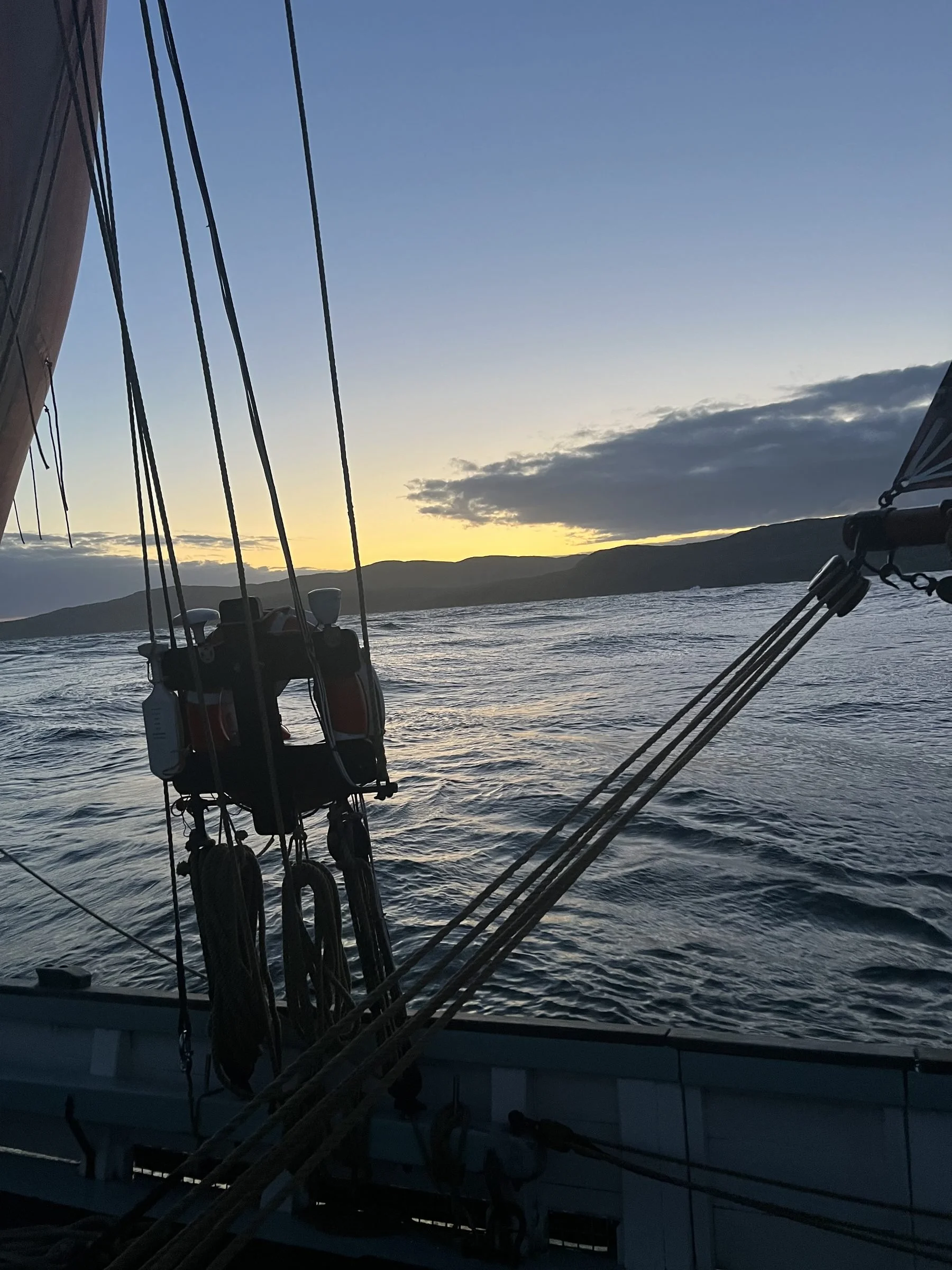 View from the sailboat, Provident,  at sunset with visible rigging and sails, ocean water, distant mountains, and a partly cloudy sky.
