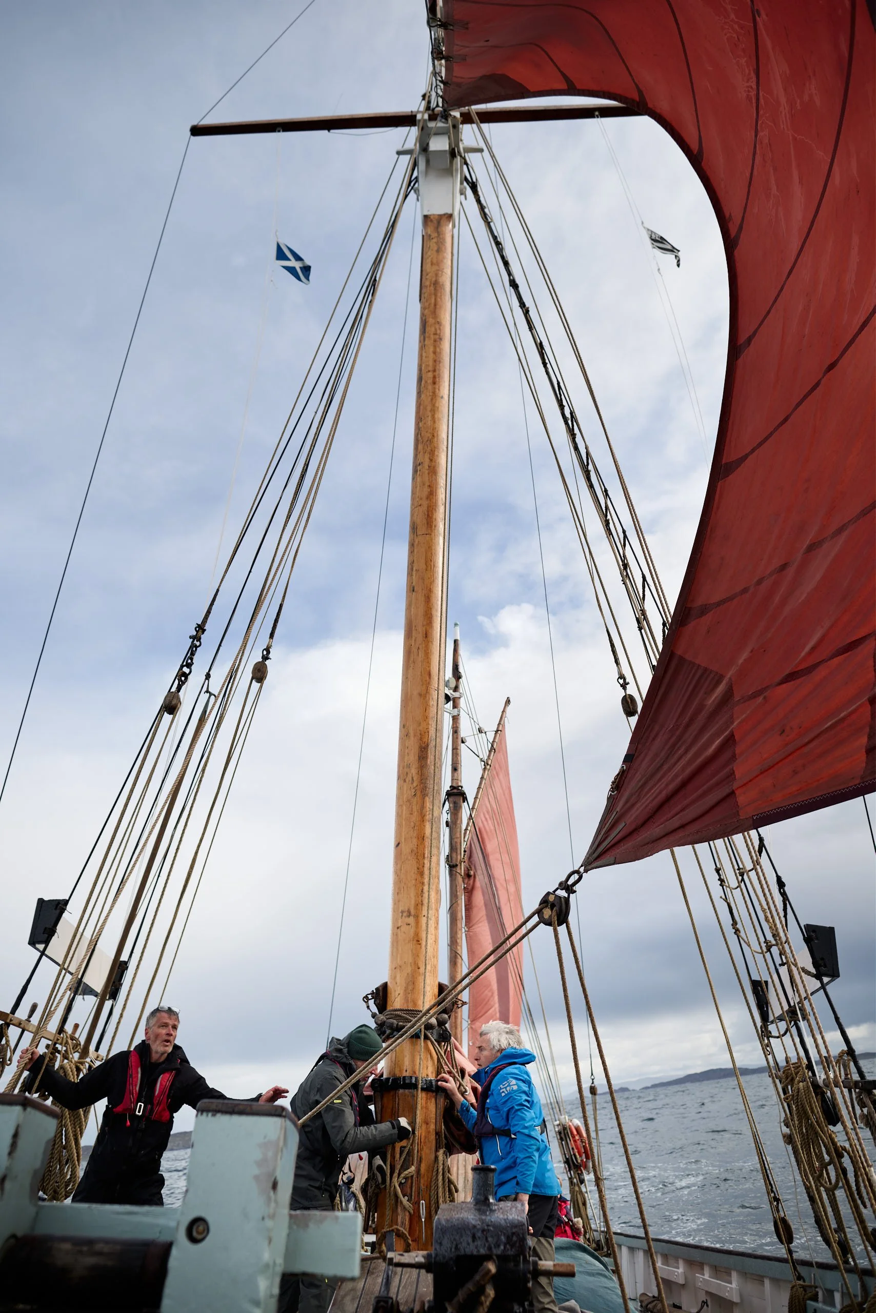 A group of people aboard a sailing ship with large red sails, standing on the deck and looking around, with the sea and a distant shoreline visible in the background.