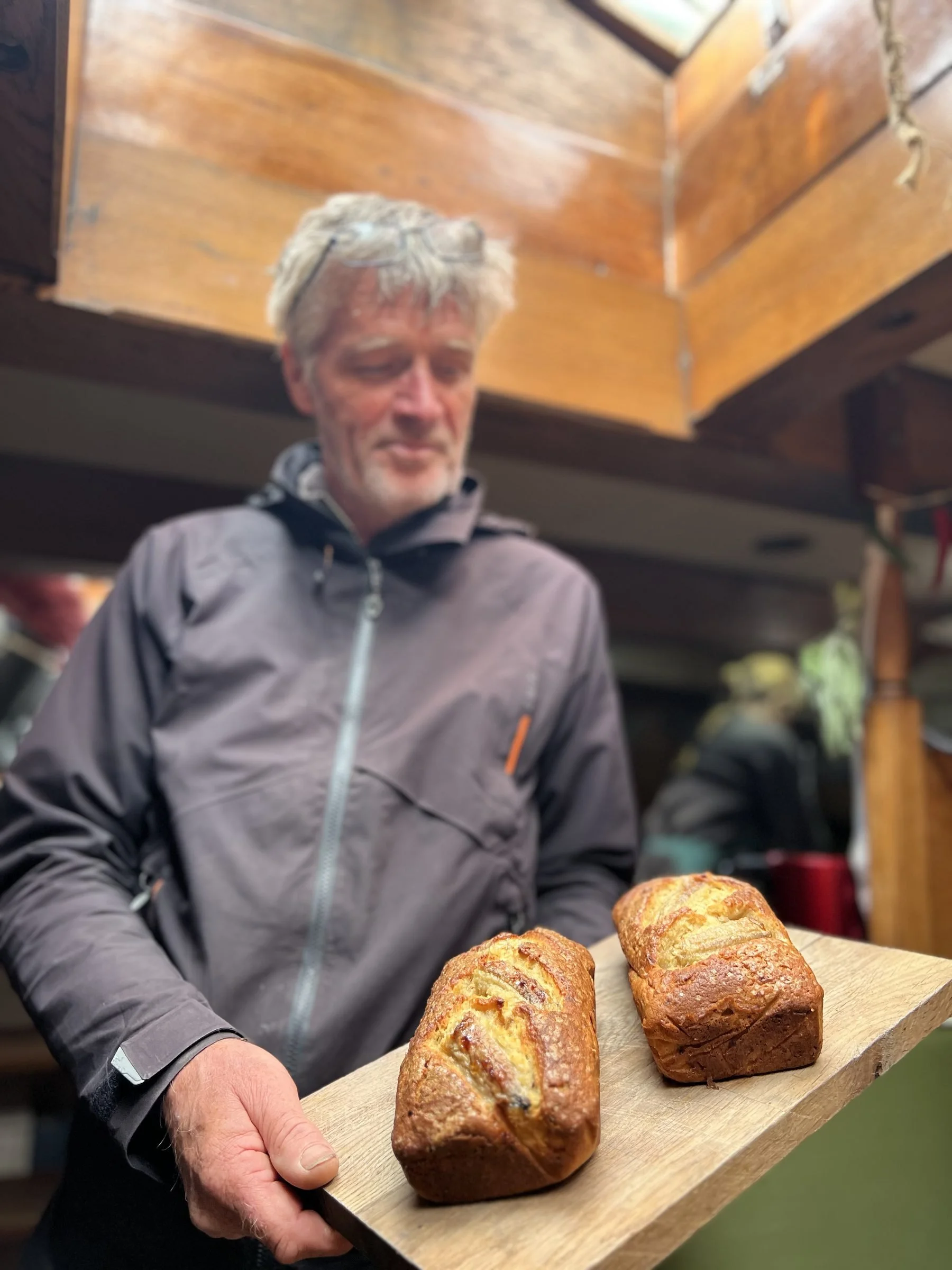 A man outdoors holding a wooden tray with three loaves of freshly baked bread, with a wooden structure and greenery in the background.