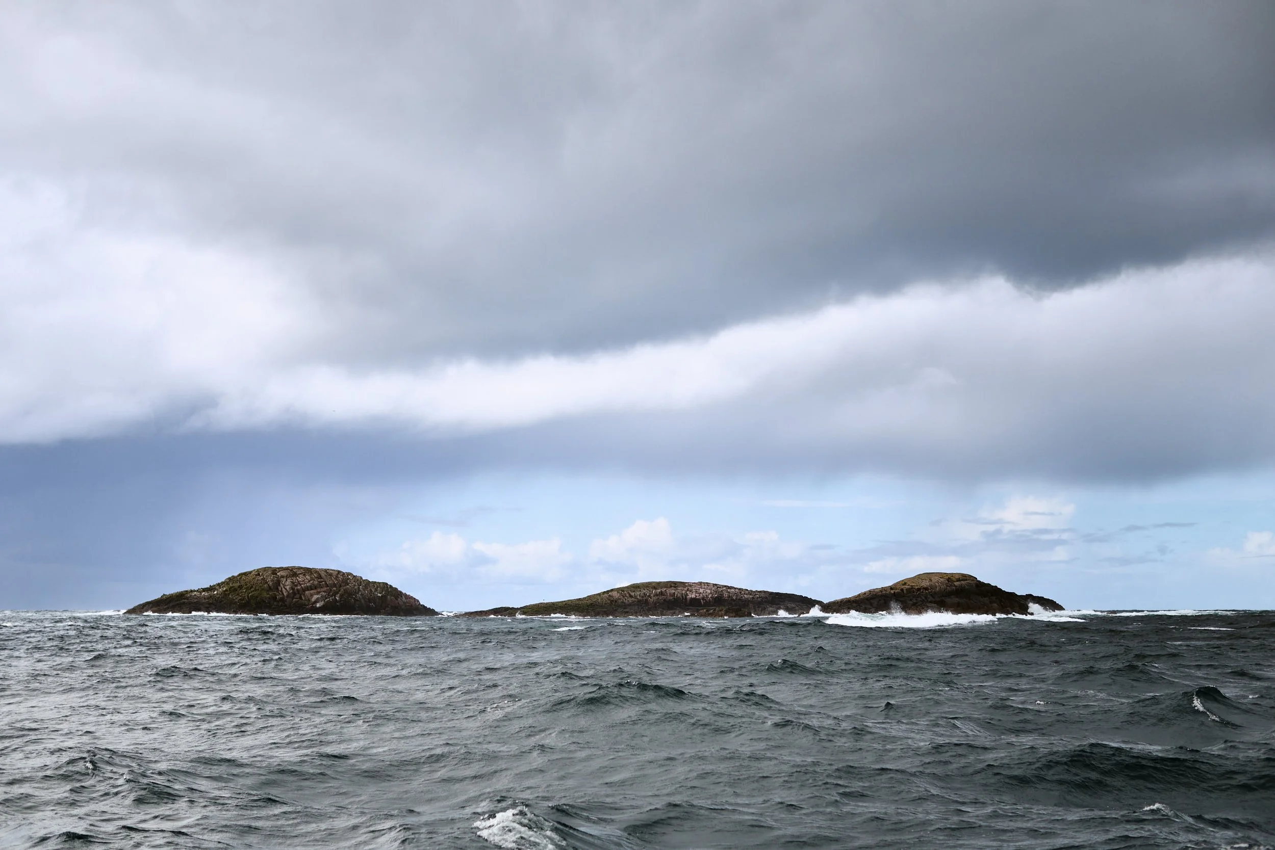 A body of water, possibly an ocean or sea, with small waves and rocks in the distance under a cloudy sky.