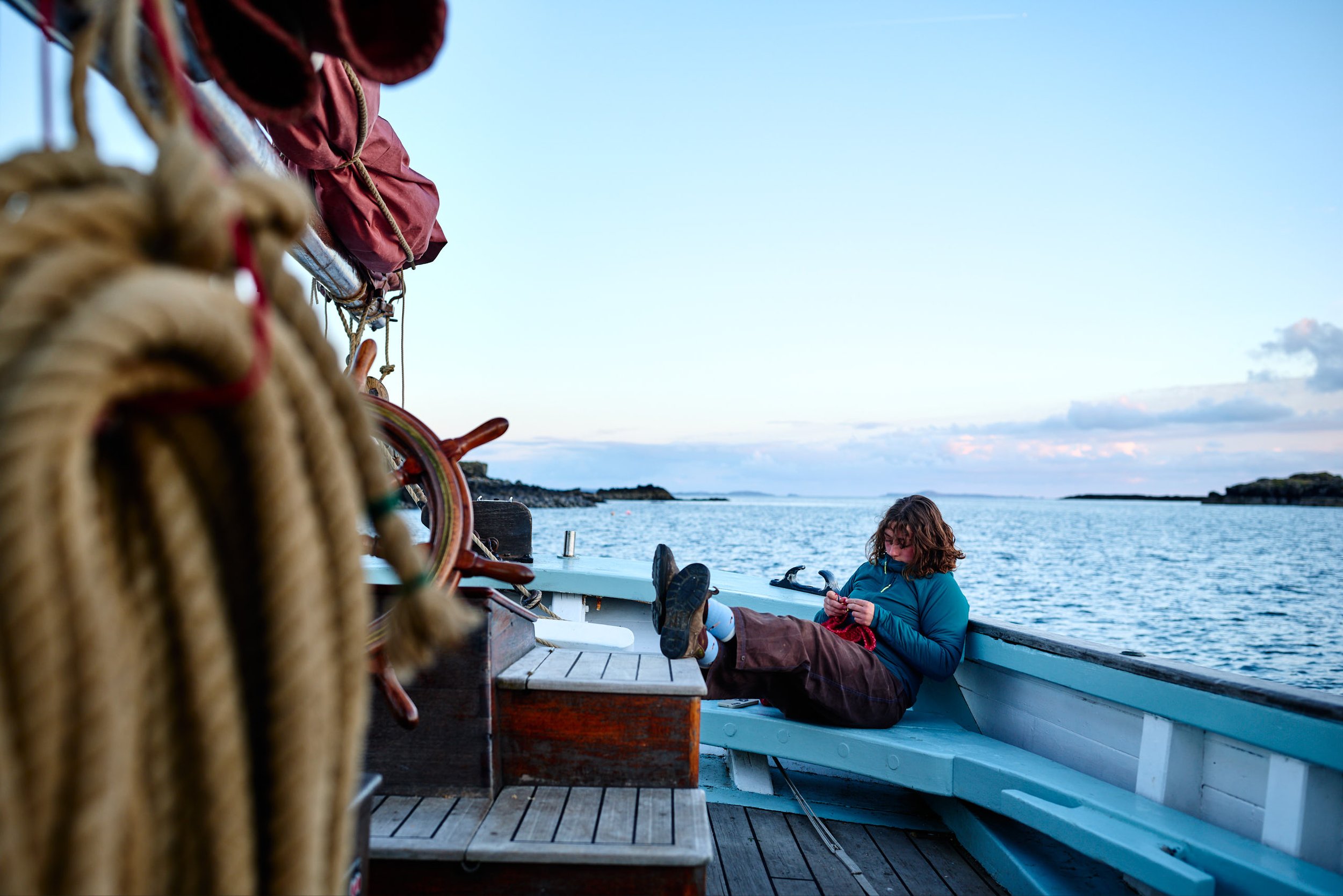 A person with curly hair relaxing on a boat, leaning back and reading a book, with a young girl sitting nearby by the water, under a blue sky with some clouds.