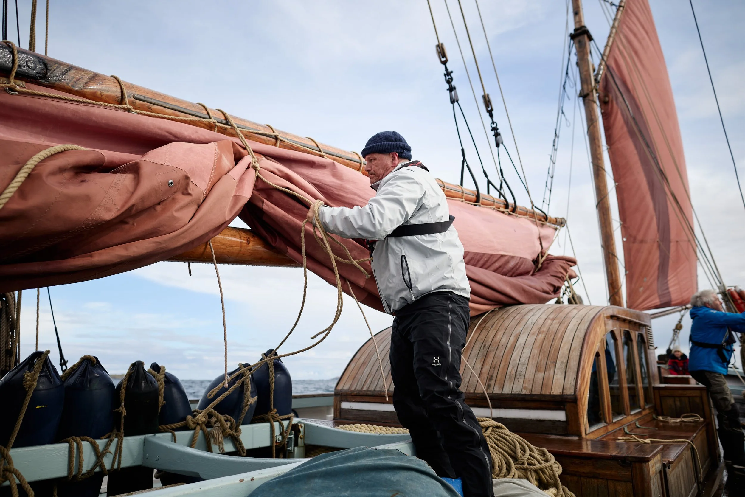 A man in a white jacket and black beanie working on the sails of a wooden sailing ship during daytime. Other crew members are visible in the background, with the sea and sky behind.