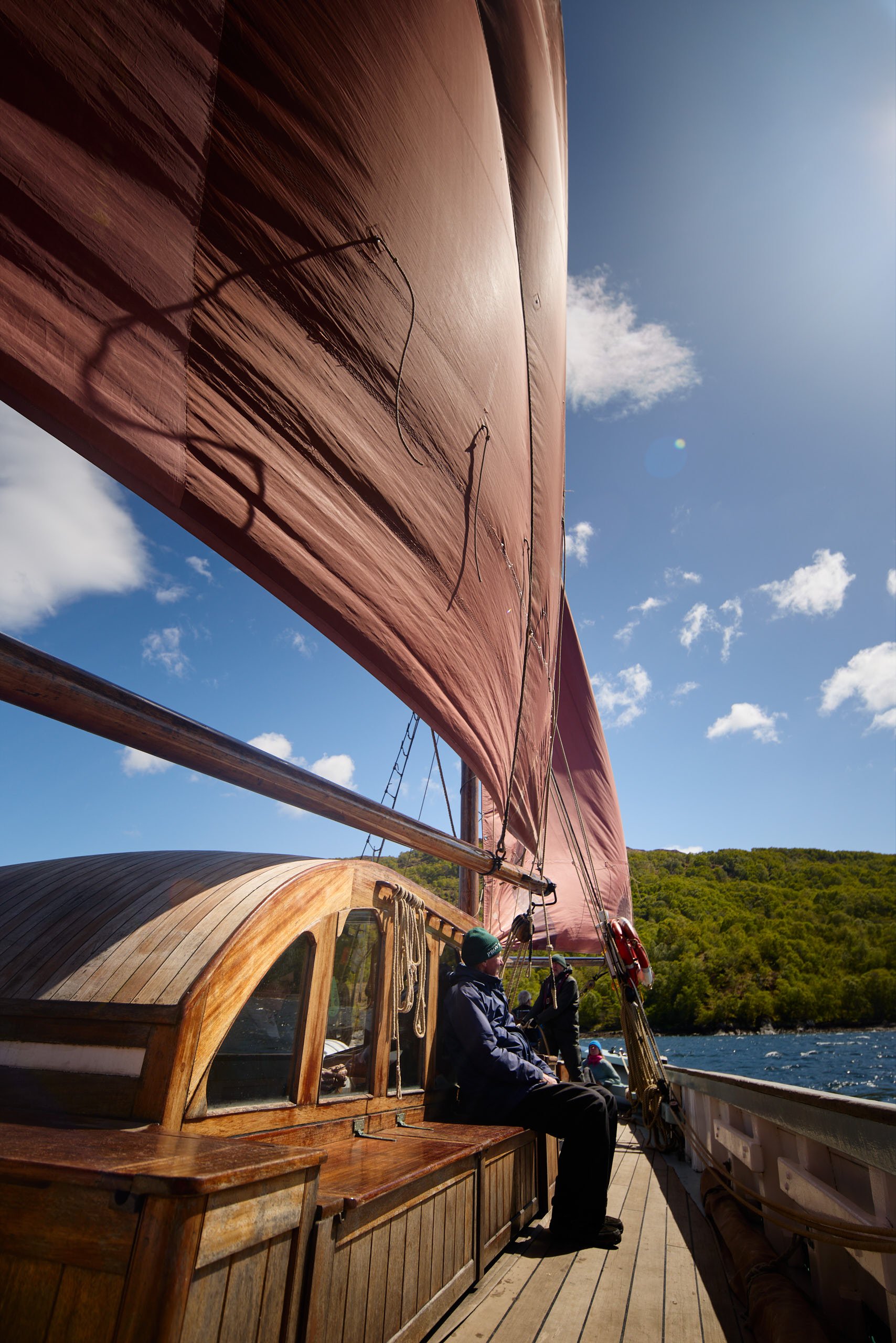 Provident with a wooden exterior and pink sails on a body of water, with people sitting and standing on the deck. The background features a green hilly landscape under a partly cloudy sky.