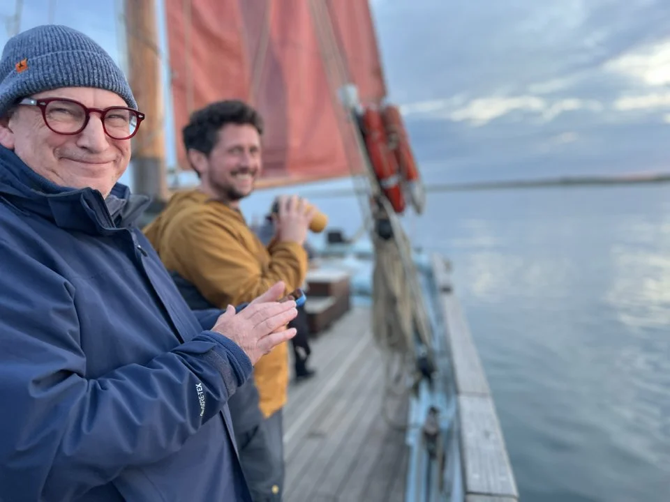 Two men standing on a sailboat deck, smiling at the camera, with water and a cloudy sky in the background.