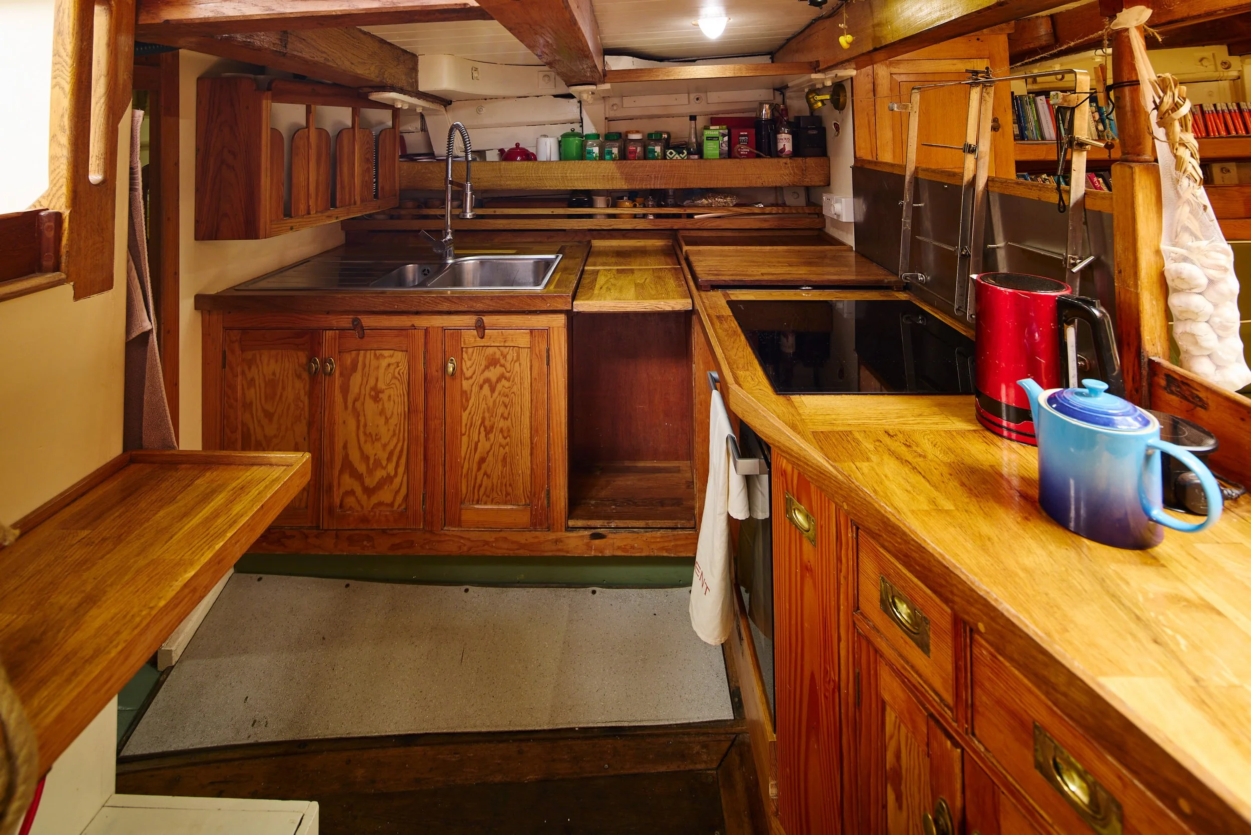 A small kitchen with wooden cabinets and countertops, a stainless steel sink, various jars and containers on open shelves, a red electric kettle, and a blue and red mug on the counter. There are open shelves with books, a ladder, and a bundle of garl