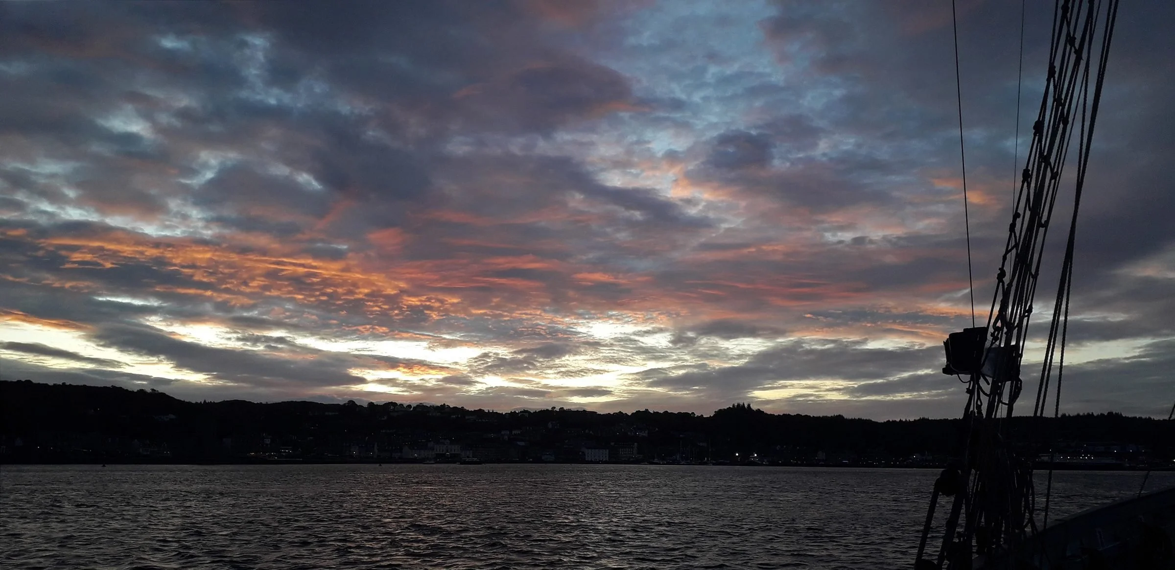 Sunset over a body of water with a cloudy sky and a silhouette of a ship's rigging on the right.