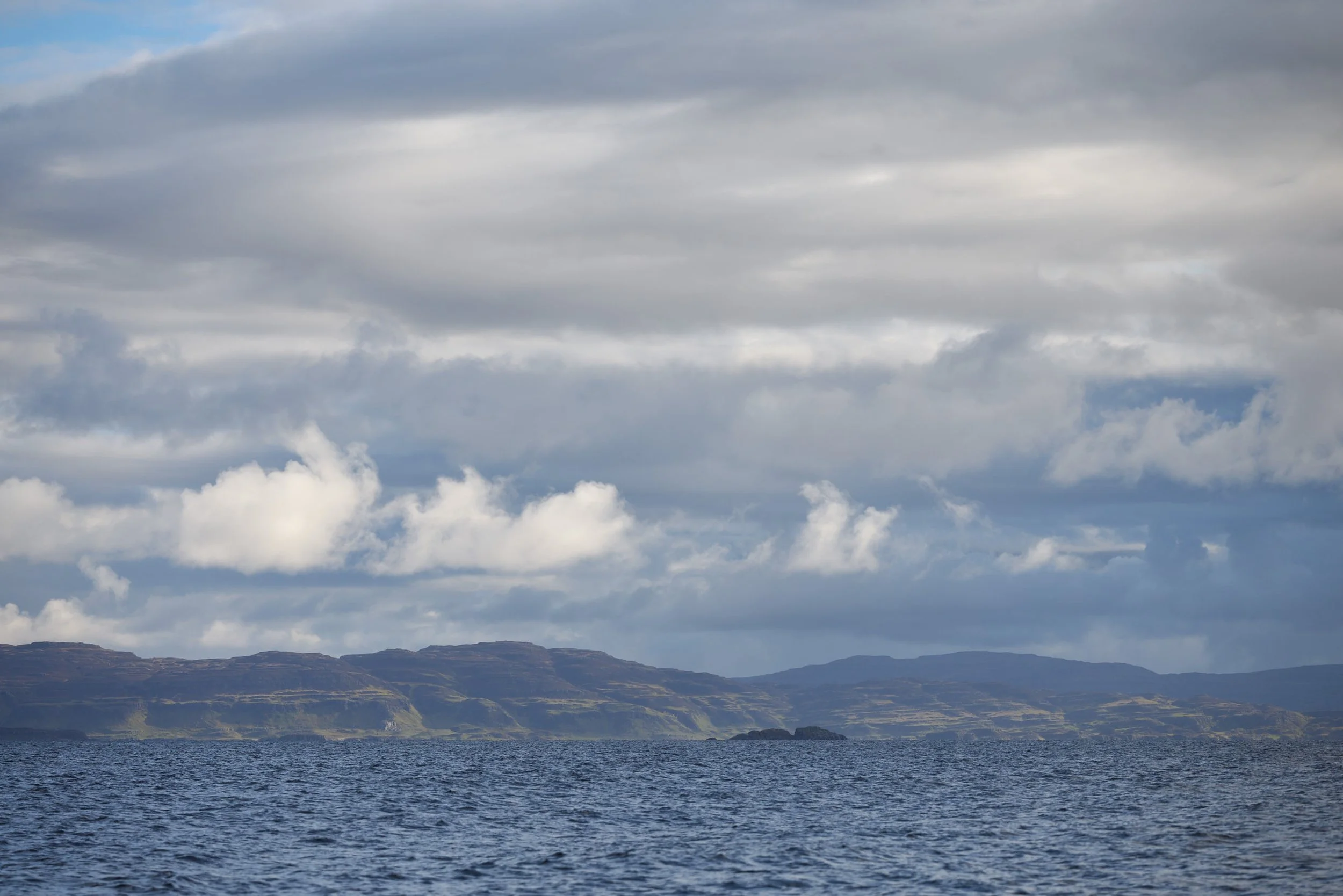 A vast body of water with waves, mountain ranges in the background, and a cloudy sky overhead.