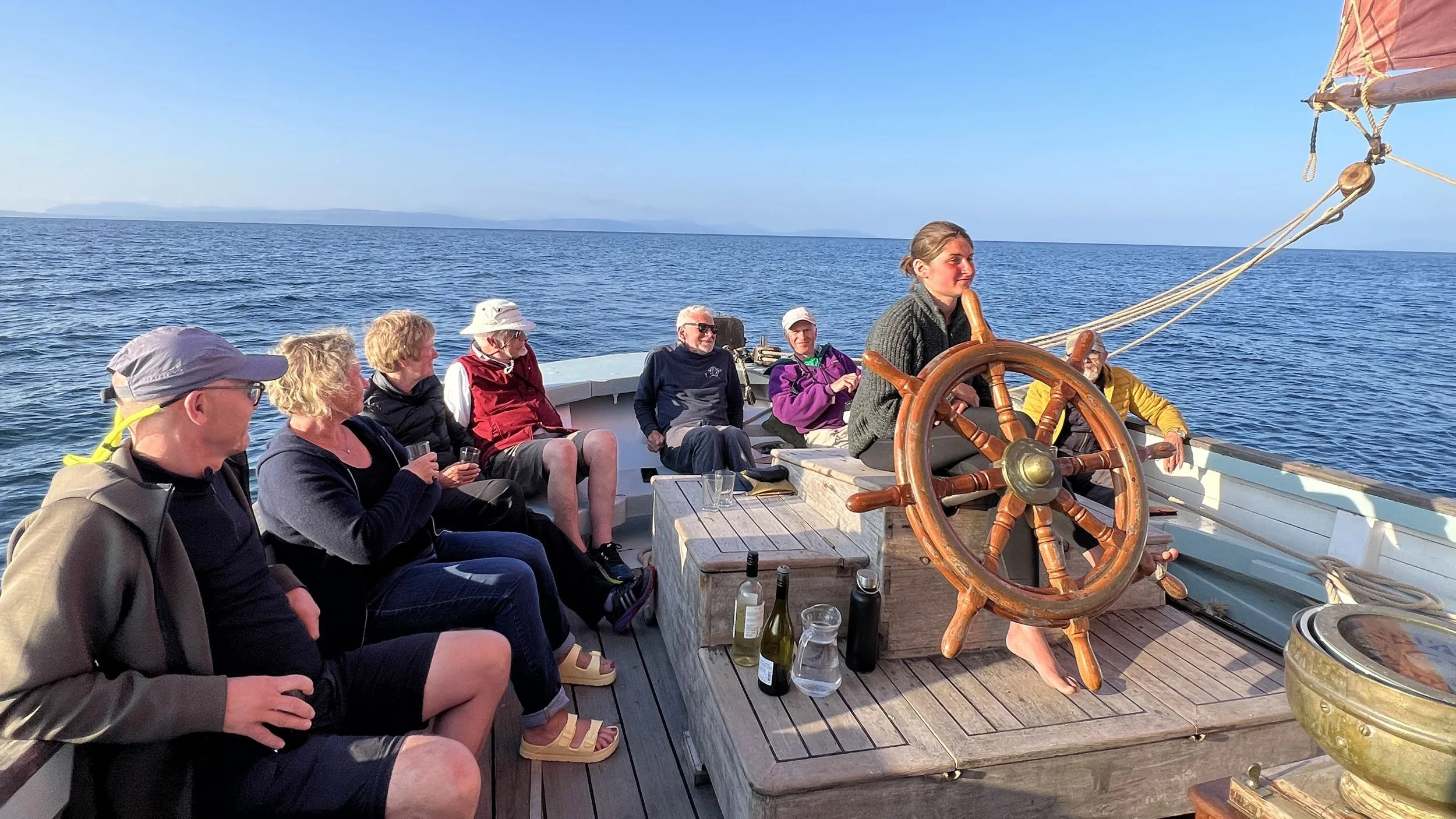 A group of people sitting on a boat, enjoying a boat ride on a large body of water with land visible in the background, with a woman at the helm steering.