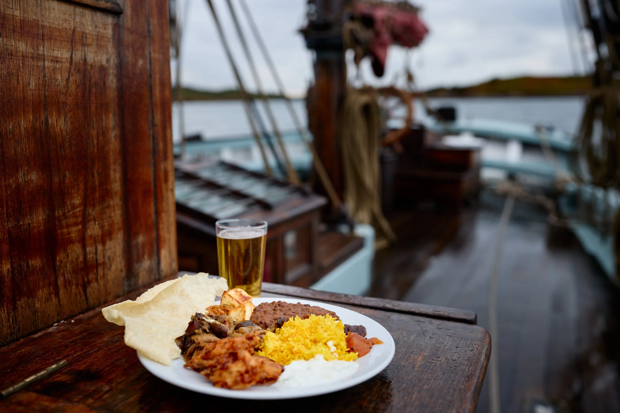 A plate of food with rice, various meats, bread, and vegetables on a wooden table outdoors near a lake, with a glass of beer in the background.