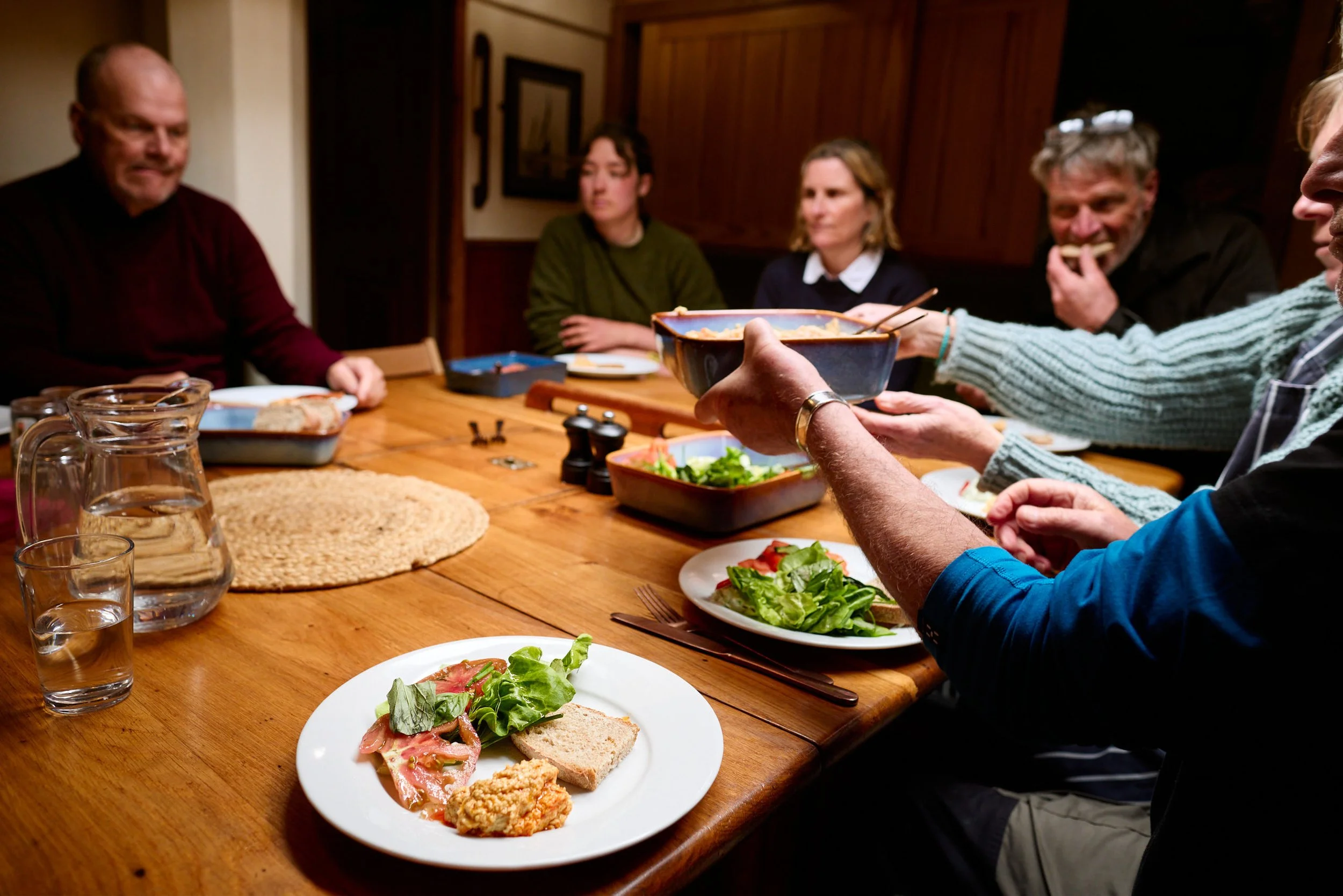 People gathered around a wooden dining table having a meal, with food on plates, including salad, bread, and fried food, in a warmly lit room with wooden paneling.
