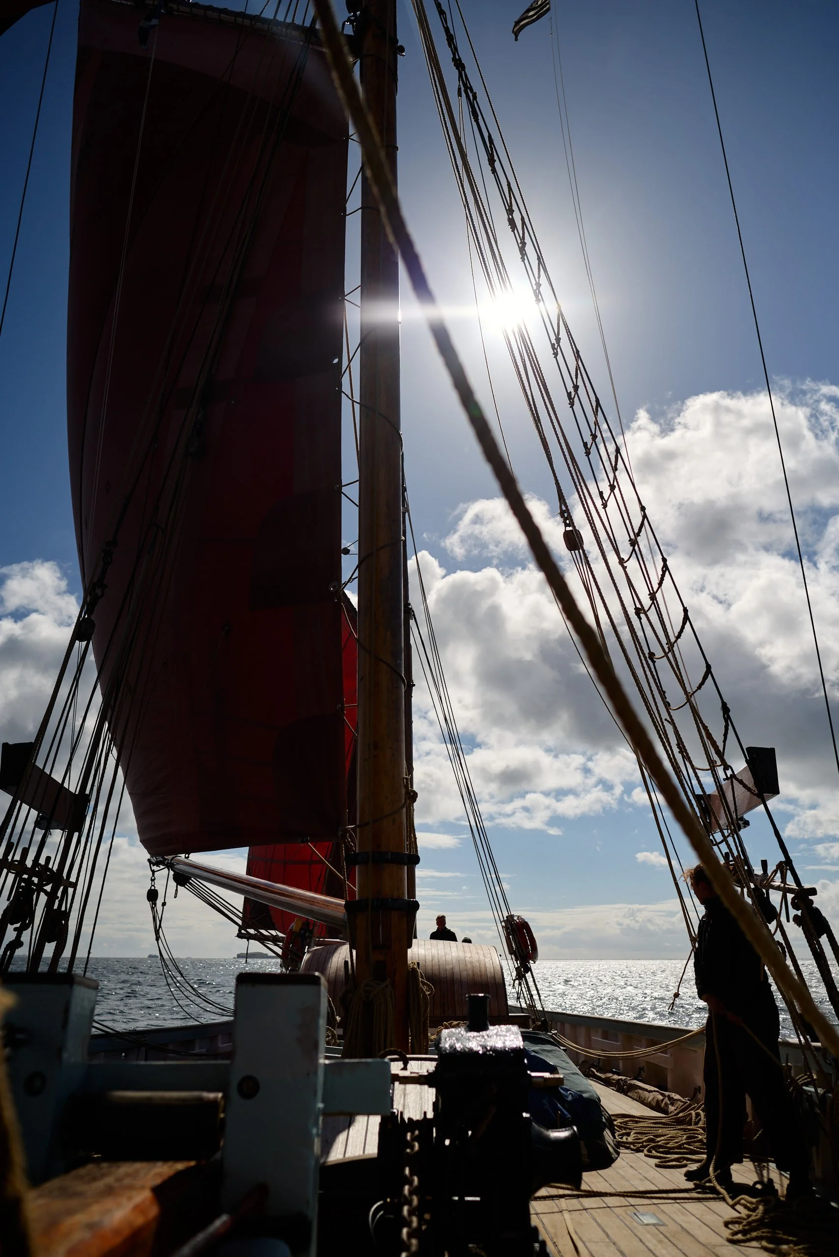 View from the deck of a sailing ship looking up at the mast and rigging with the sun shining behind, sea and sky visible in the background.
